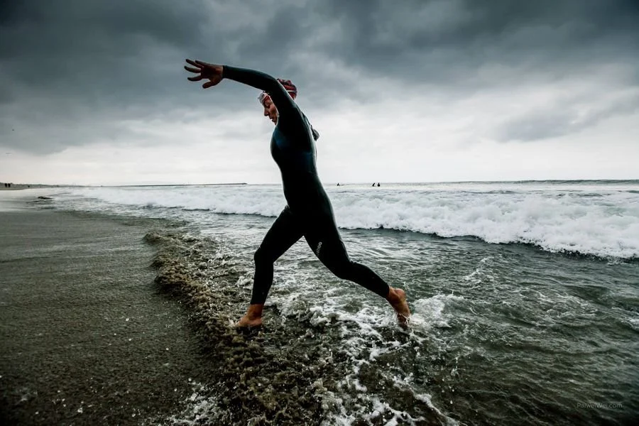 Person in a wetsuit balancing on the edge of the ocean in a surfing pose during cloudy weather.