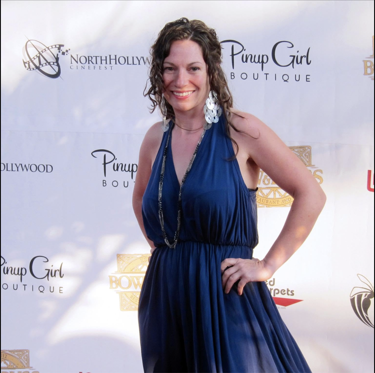 A woman with curly brown hair and light skin, wearing a sleeveless blue dress and large dangling earrings, standing with her hand on her hip and smiling at a red carpet event.