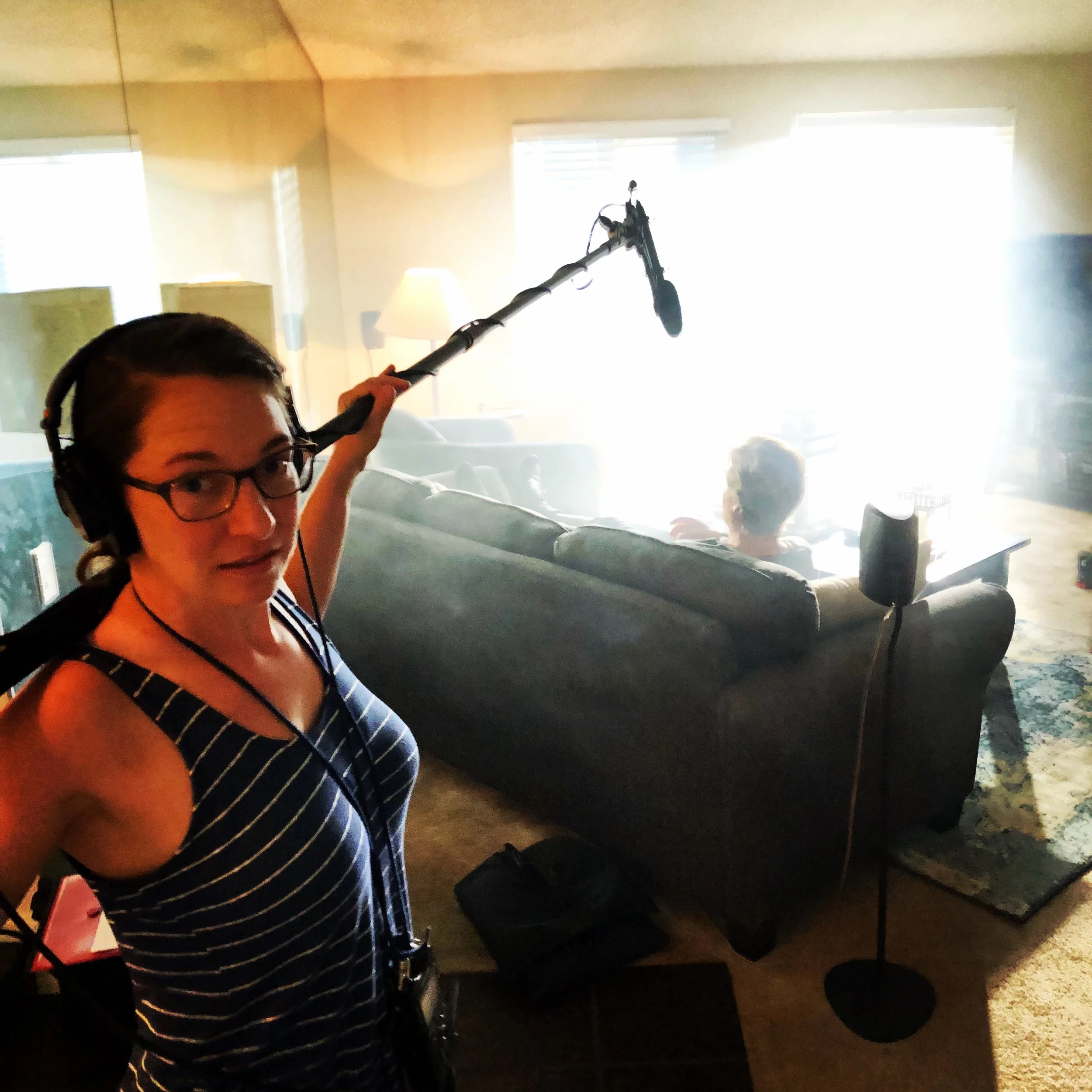 A person with glasses and dark hair holding a boom microphone in a room with bright windows, a lamp, and a motorcycle in the background.