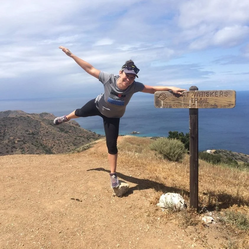 Woman in athletic clothing and cap balancing on one leg next to a trail sign on a hilltop with ocean view in the background.