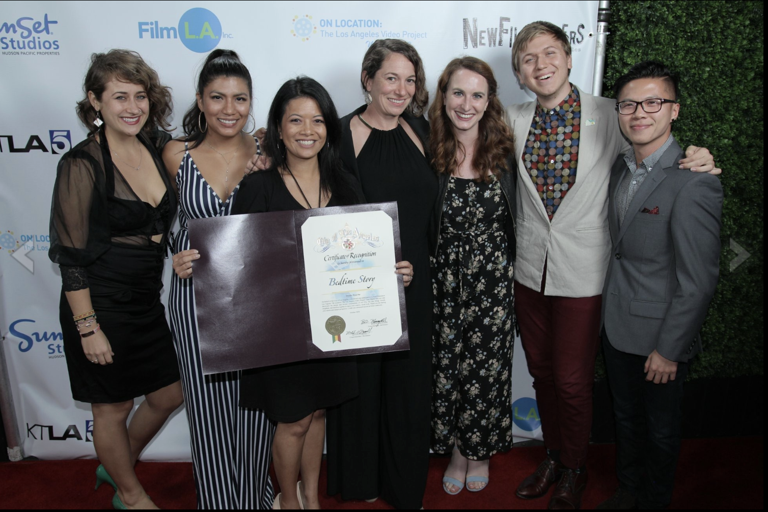 Group of seven diverse individuals posing at an event, with one holding a certificate of recognition. They are dressed in formal and semi-formal attire, standing on a red carpet against a backdrop with logos and writing related to a film or media pro