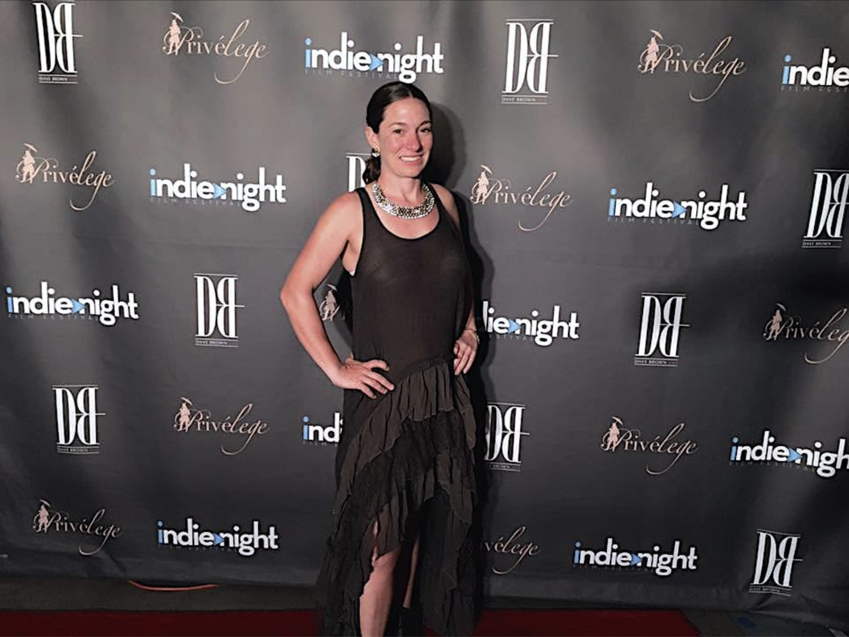 A woman standing on a red carpet at a black-themed event backdrop with logos, smiling, wearing a black sheer sleeveless dress with layered ruffles and jewelry.