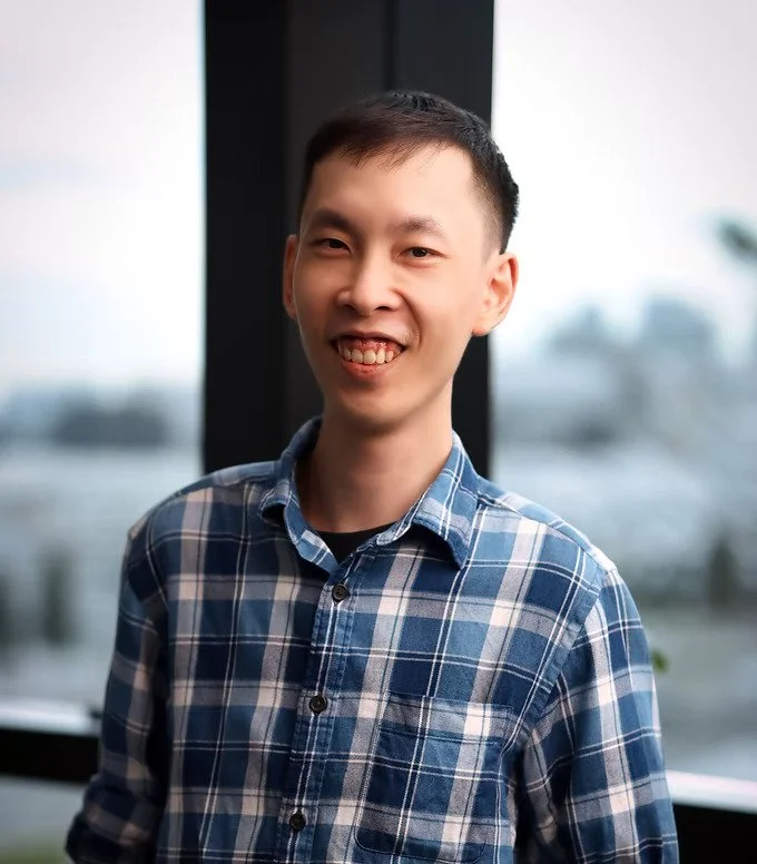 A young man with short black hair, smiling, wearing a blue plaid shirt, standing indoors near a window with a blurred outdoor view.