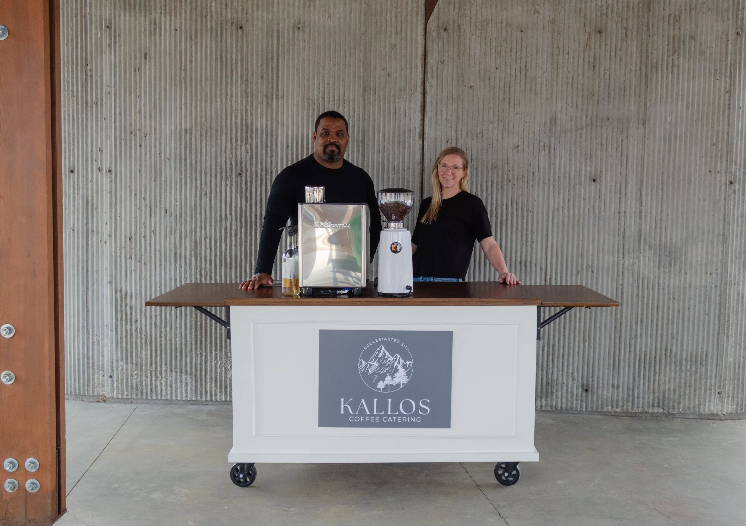 Two people standing behind a white coffee cart with a Kallos Coffee Catering logo, set against a concrete wall, with coffee brewing equipment on the cart.