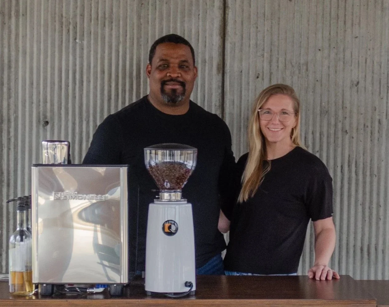 A man and woman standing behind a coffee grinder and bottles on a wooden table, smiling at the camera against a corrugated metal wall.