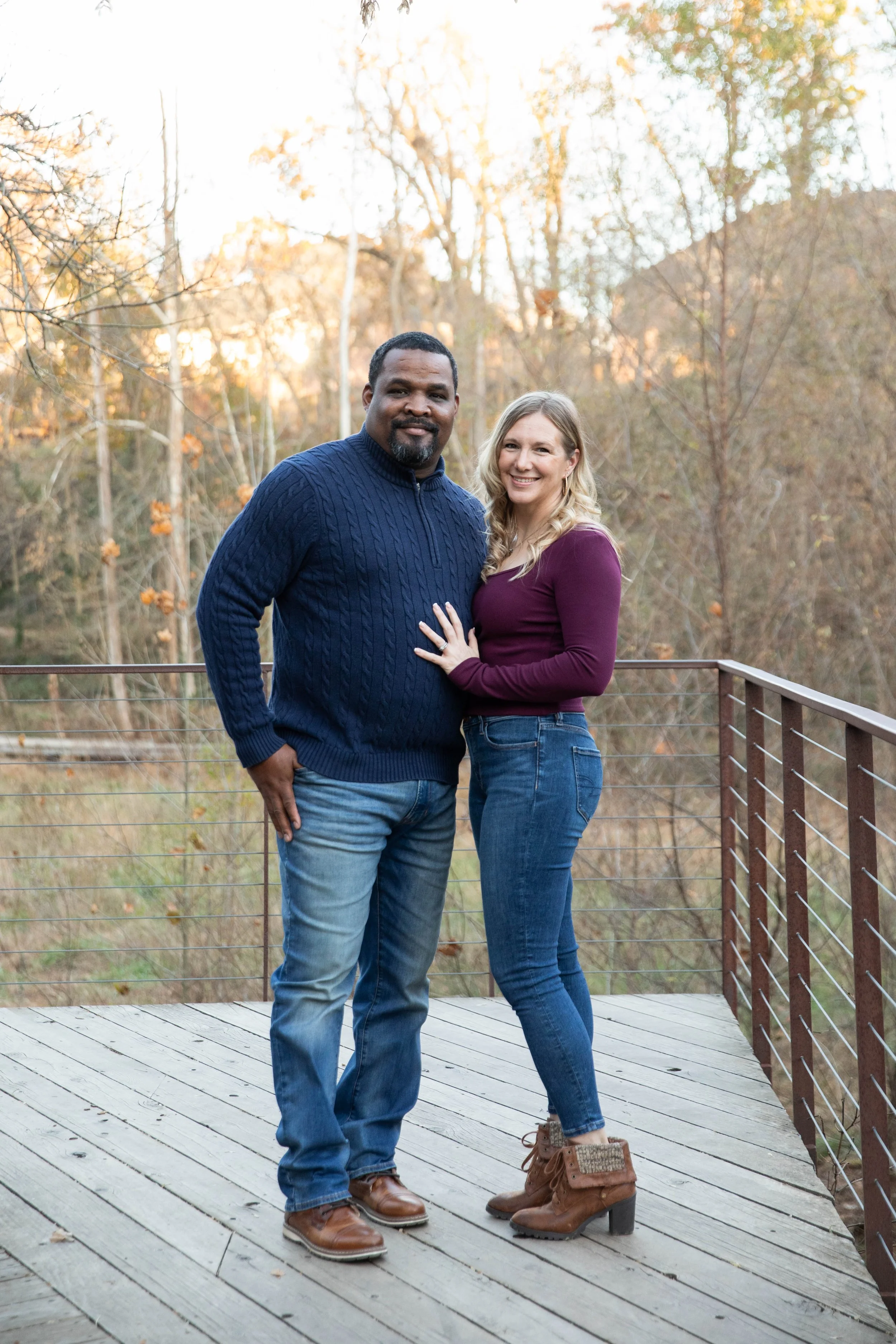 A couple standing on a wooden deck outdoors during fall, with trees in the background. The man is wearing a navy blue sweater and jeans, and the woman is wearing a burgundy long sleeve top, jeans, and brown boots. They are smiling and standing close together, with the woman resting her hand on the man's chest.