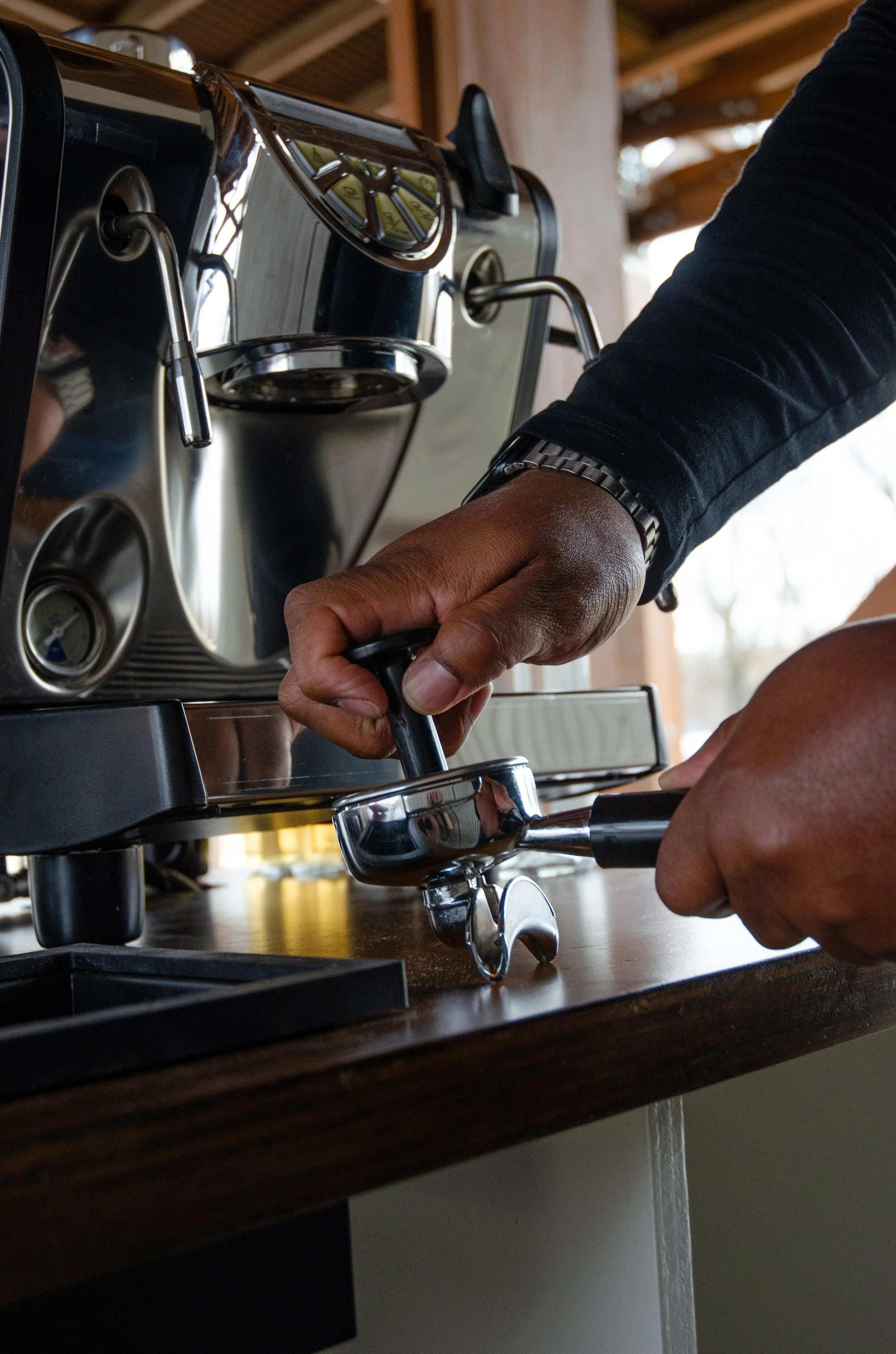 A person tamping coffee grounds in a portafilter of a commercial espresso machine. locally owned coffee bar