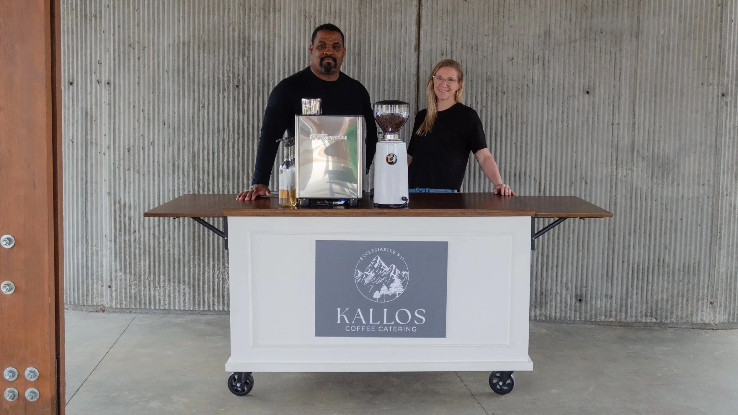 Two people standing behind a coffee catering cart with Kallos Coffee Catering logo, one man with dark skin and beard, and one woman with fair skin and blonde hair, in front of a textured concrete wall. locally owned mobile coffee catering.