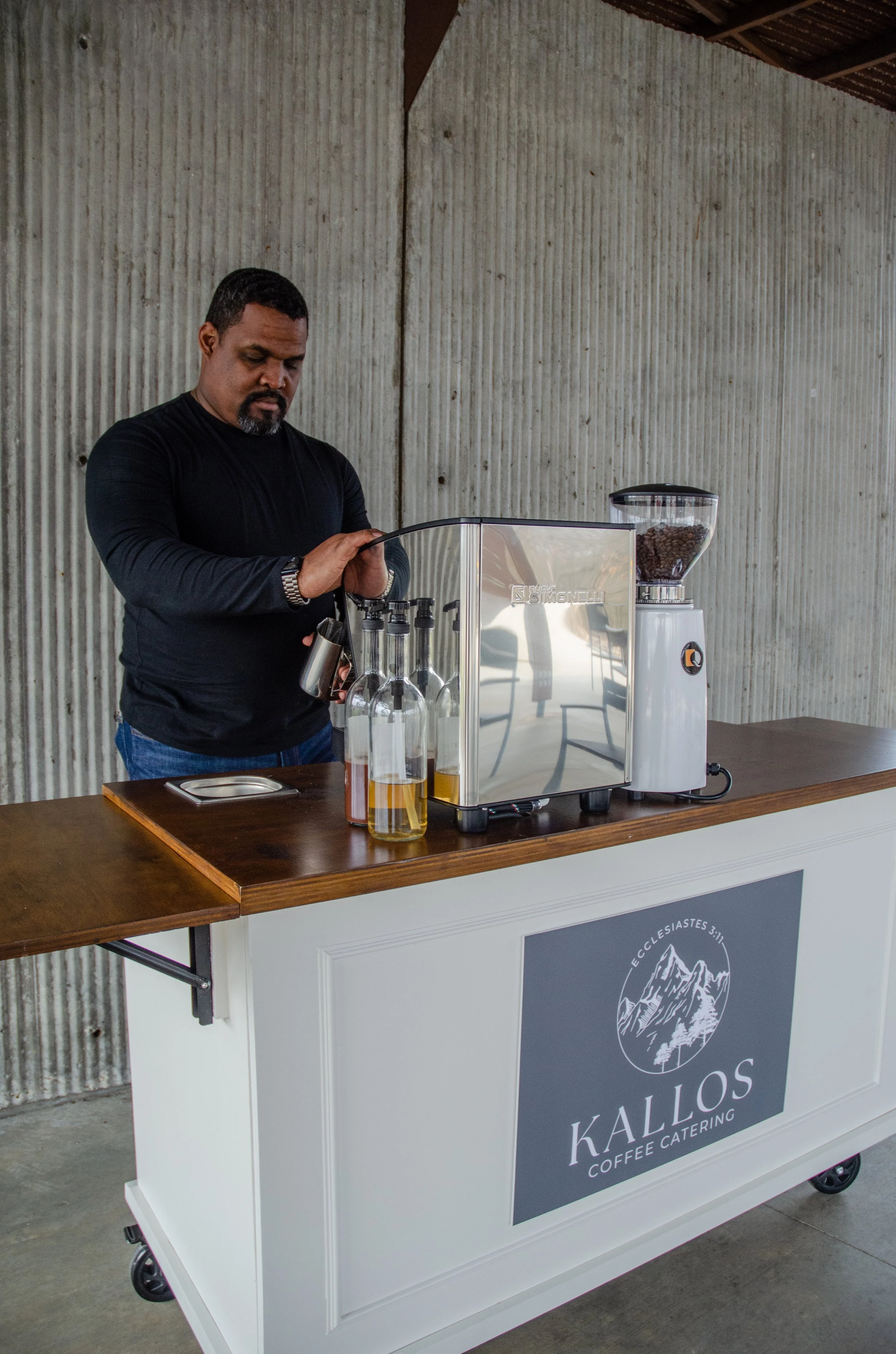 Man in black shirt preparing coffee using a coffee machine on a wooden cart labeled "Kallos Coffee Catering." locally owned mobile coffee catering