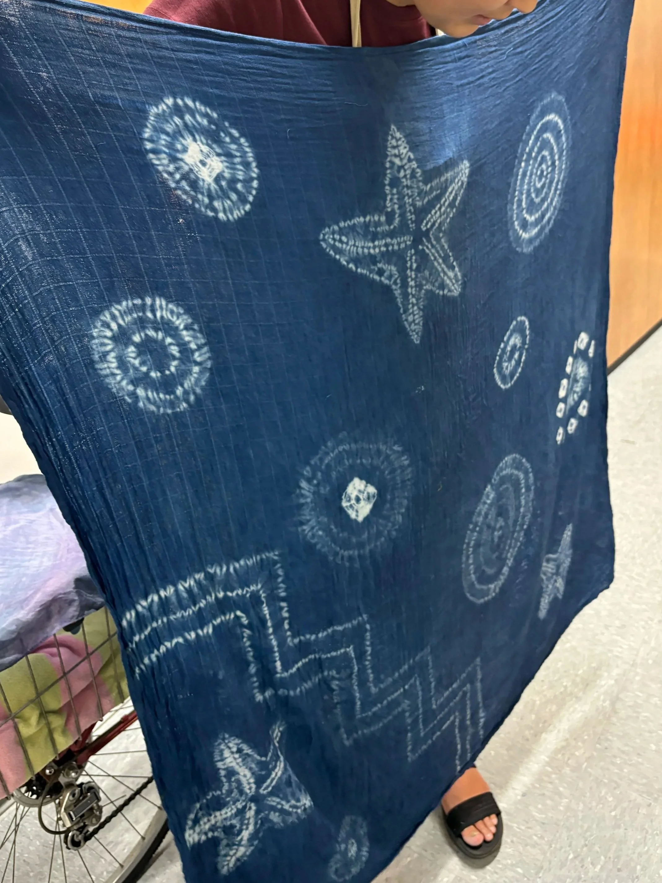 A large blue square fabric, with designs in white, created by resists during dyeing.  Circles, a zigzag, three starfish.