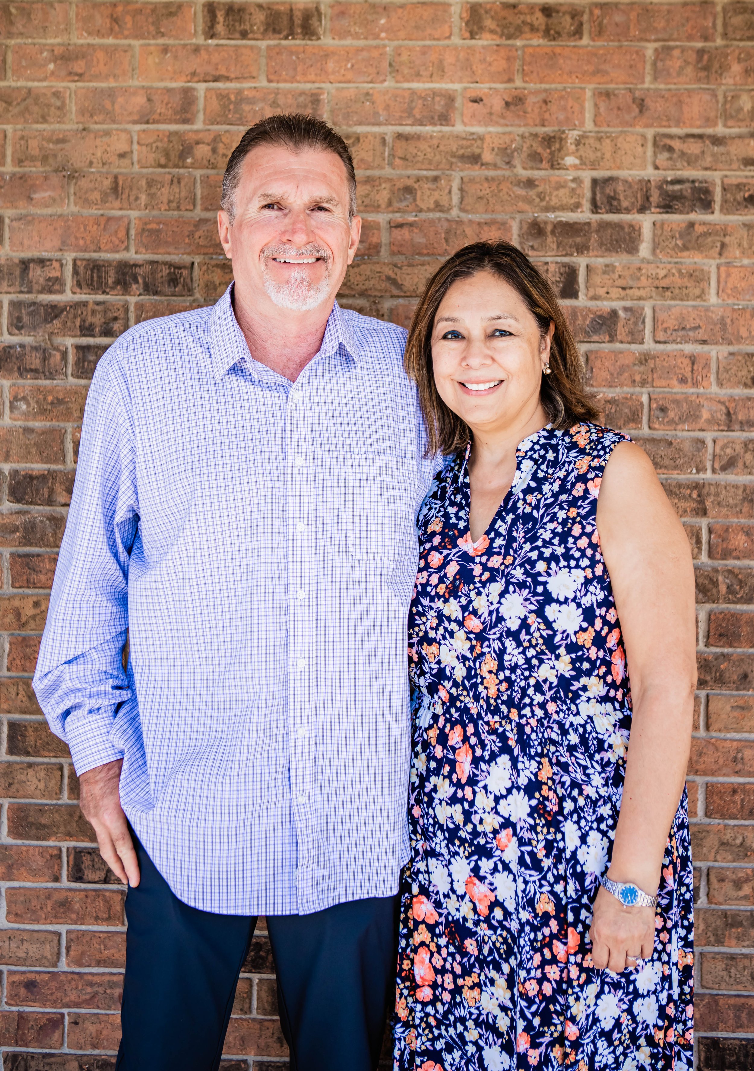 The worship leaders at New Foundation in Azle, Texas. A man and a woman standing in front of a brick wall, smiling, dressed in casual business attire, with the man wearing a light blue checkered shirt and the woman wearing a sleeveless floral dress.