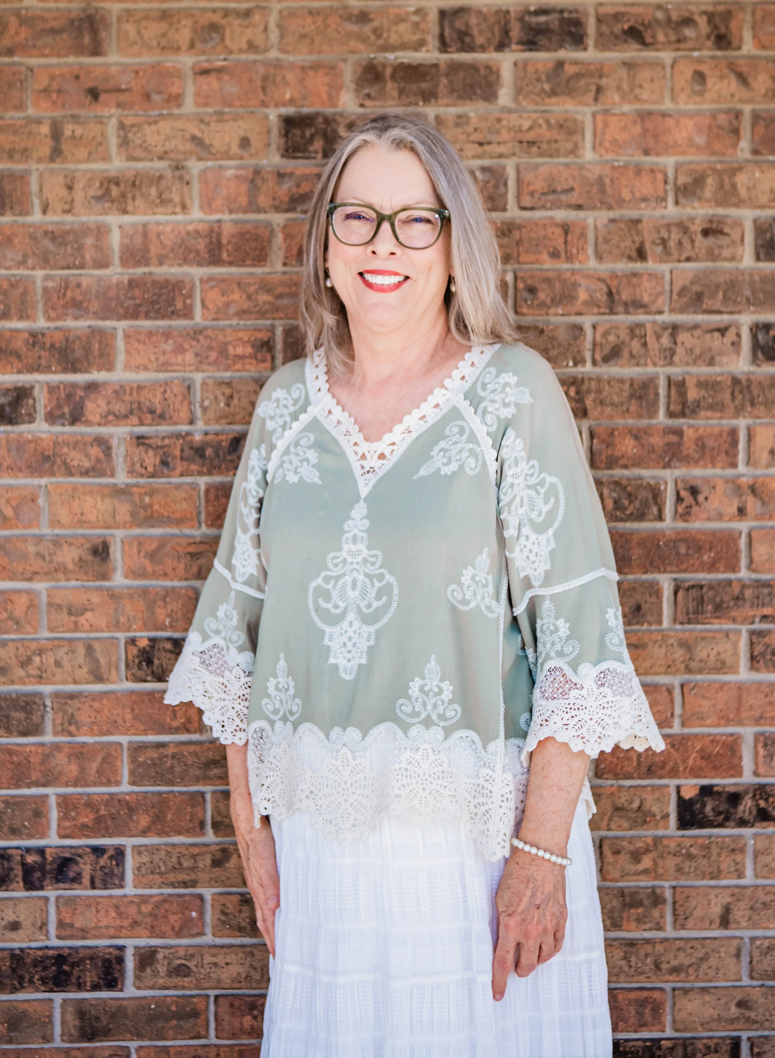 The administrative assistant at New Foundation in Azle, Texas. A smiling elderly woman with glasses and grey hair, wearing a light green and white embroidered dress, standing against a brick wall.