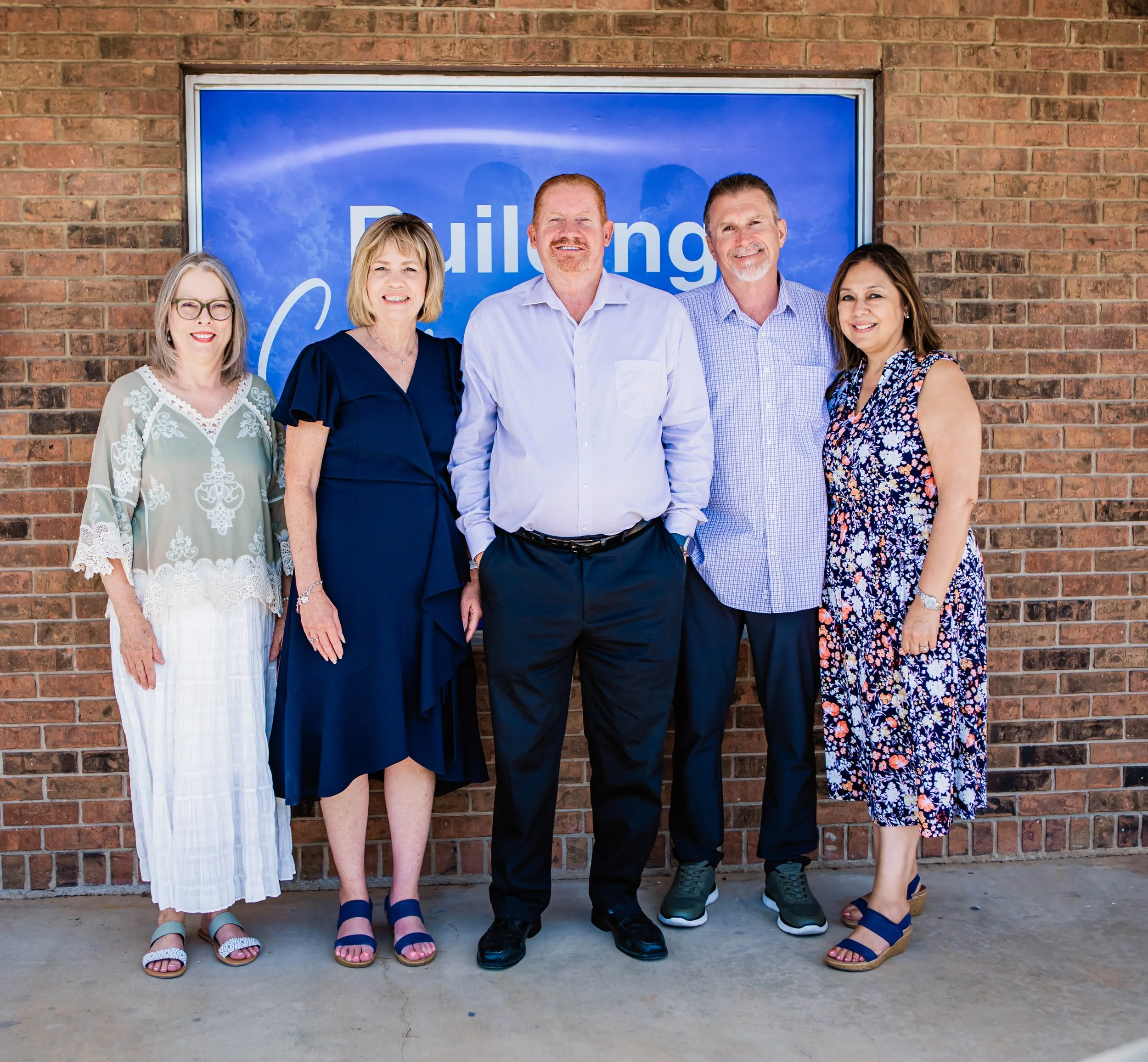 The leaders at New Foundation in Azle, Texas. A group of five people standing outside in front of a brick wall with a large blue sign, smiling at the camera.