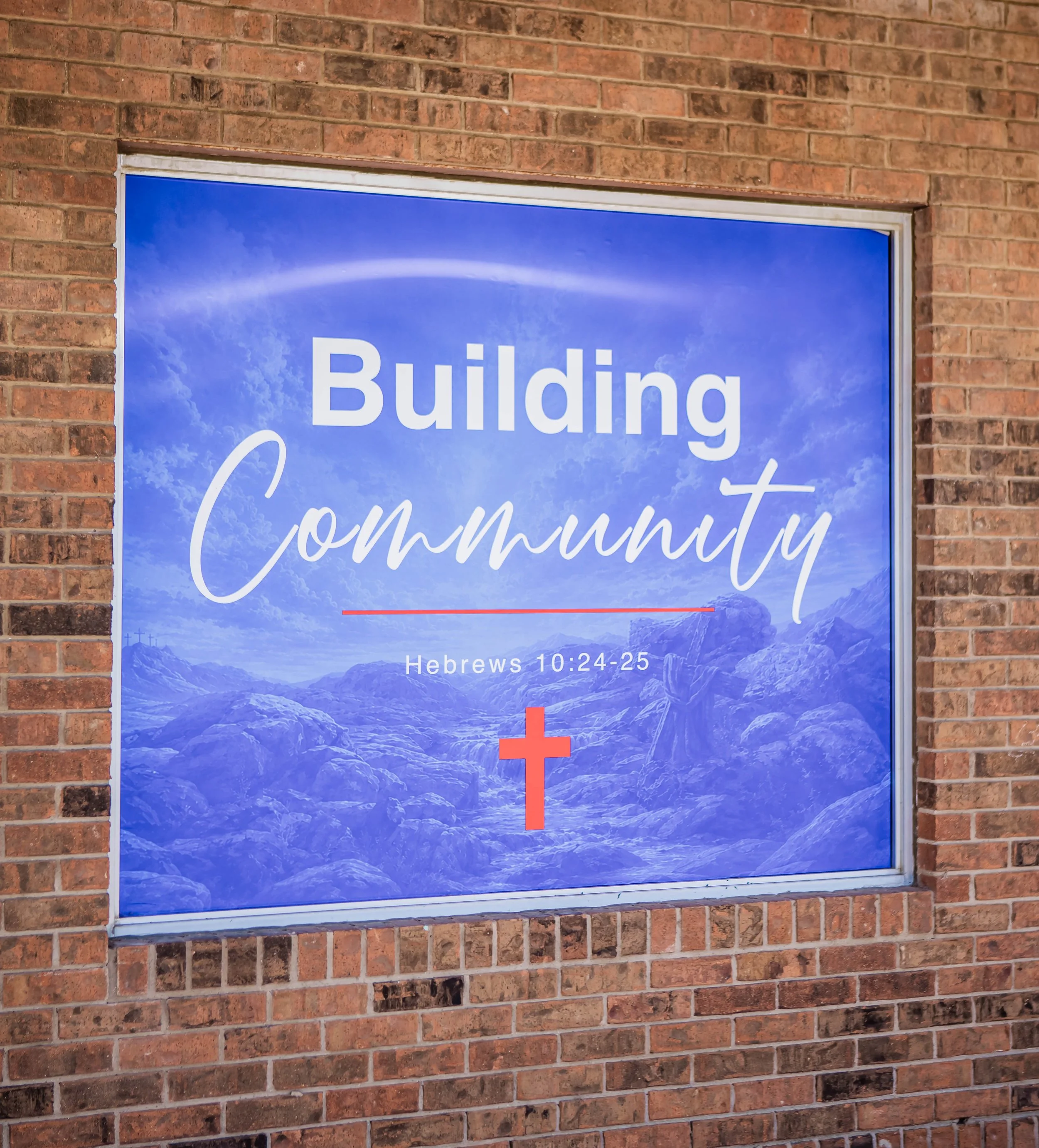 A sign on a brick wall with a mountain landscape background, displaying the words 'Building Community' and a Bible verse reference 'Hebrews 10:24-25', with a red cross symbol at the bottom. A core belief at New Foundation in Azle, Texas.