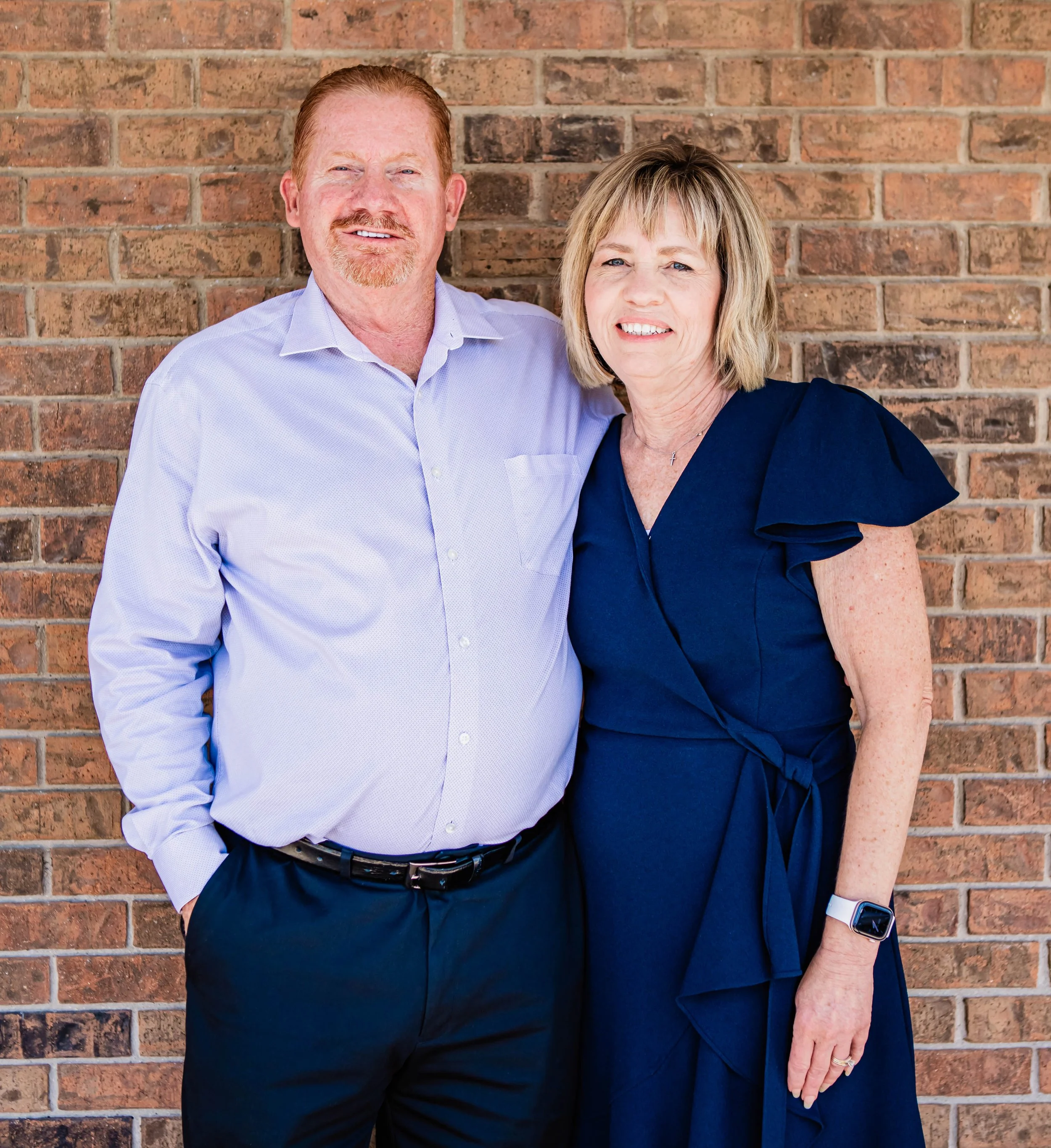 A middle-aged man with red hair and a beard wearing a light blue dress shirt stands next to a middle-aged woman with blonde hair in a navy blue dress. They are standing against a red brick wall.