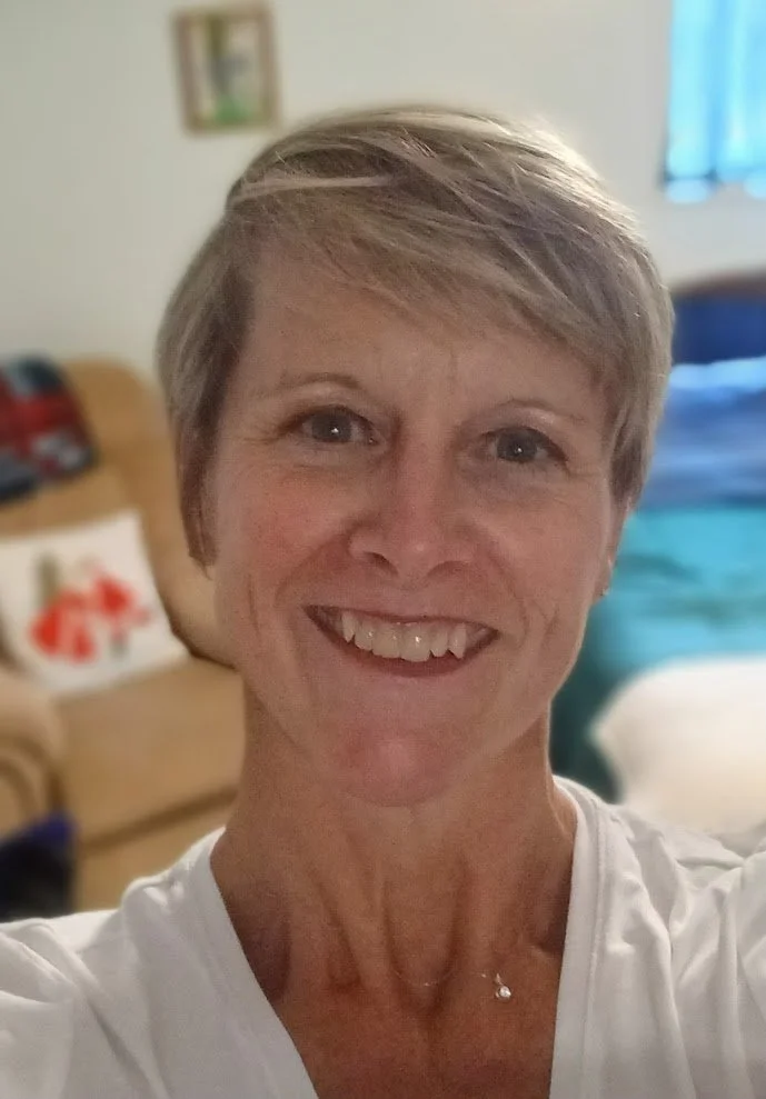 A woman with short blonde hair smiling in a cozy living room with a beige couch and blue curtains.