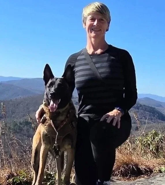 A woman kneeling outdoors with a smiling black and tan dog in front of a mountain landscape under a clear blue sky.