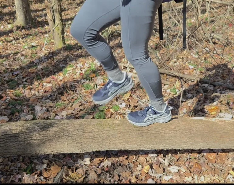 Person walking across a fallen tree trunk in a wooded outdoor area, wearing gray leggings and athletic shoes.