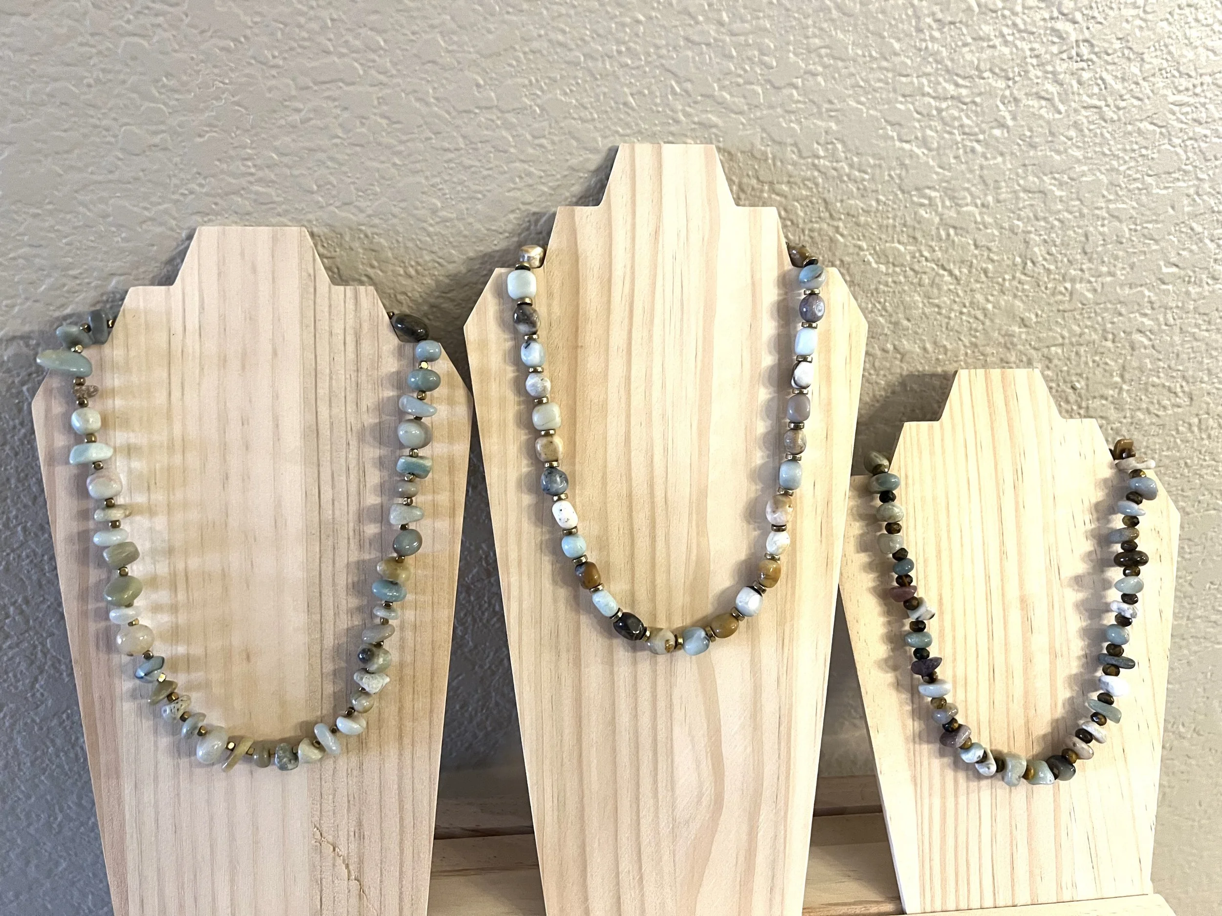 Three beaded necklaces displayed on light wooden stands against a beige wall.
