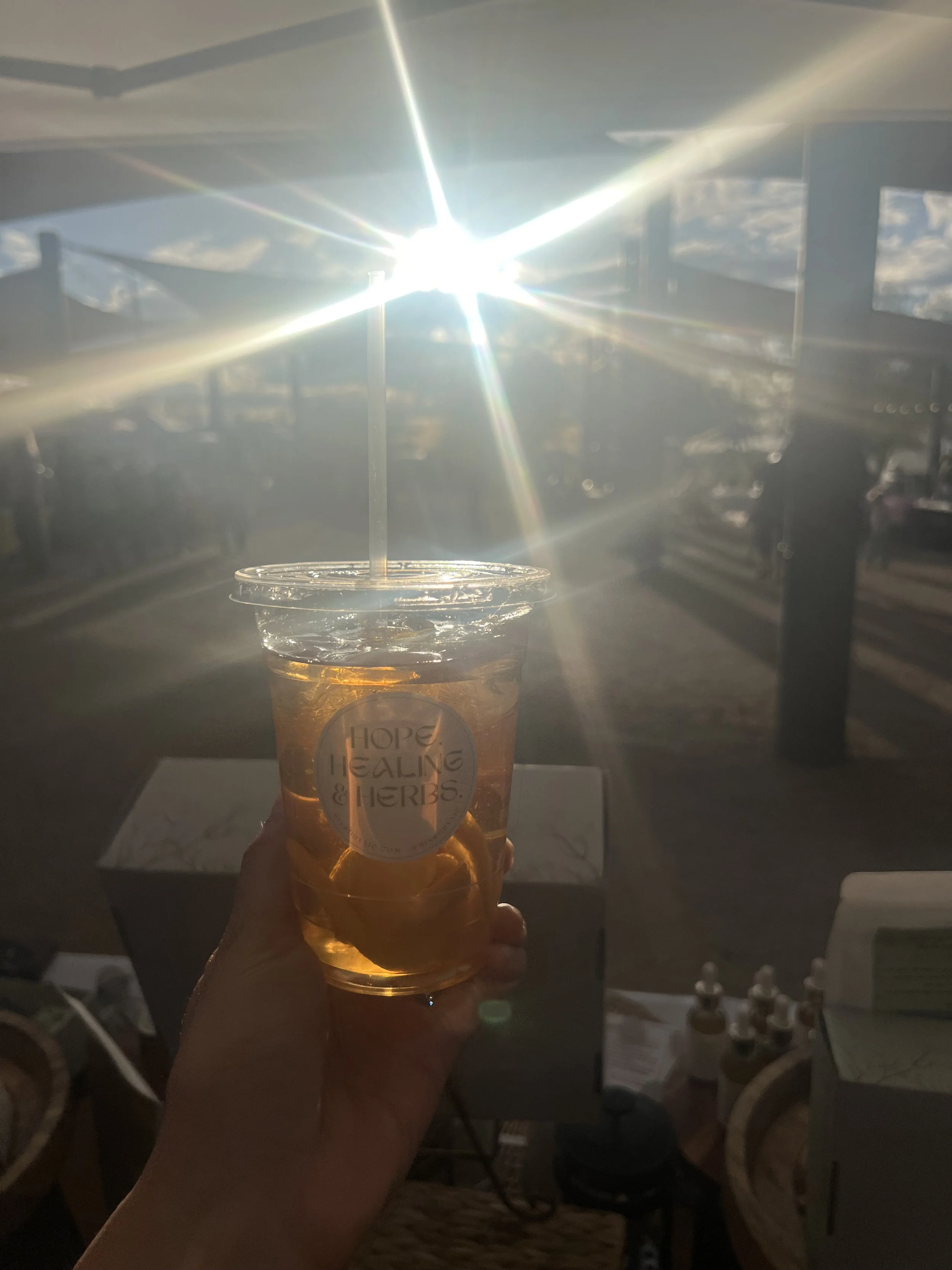 Person holding a plastic cup with iced tea and a straw, labeled 'Hope, Healing & Herbs,' with the sun shining brightly in the background and a deck in an outdoor setting.
