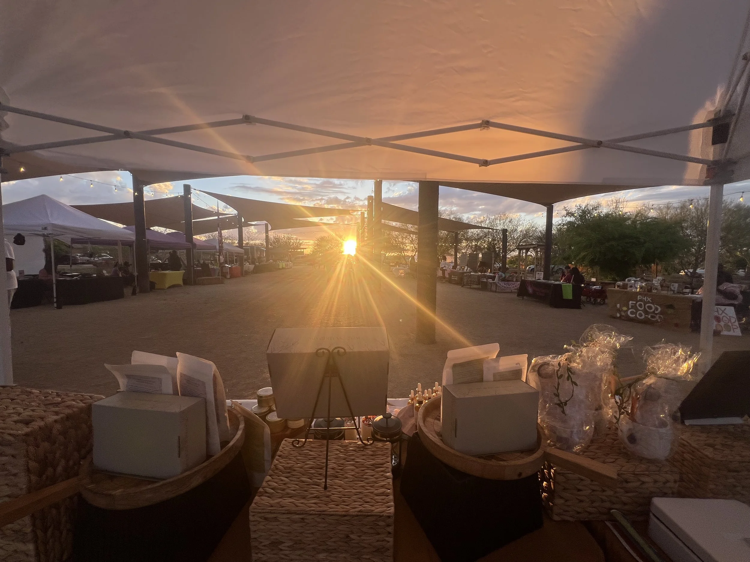 A sunset view from underneath a large canopy at an outdoor event. Several vendor booths with tables and tents are visible in the background, with people setting up or browsing. The scene has a warm, golden glow from the setting sun.