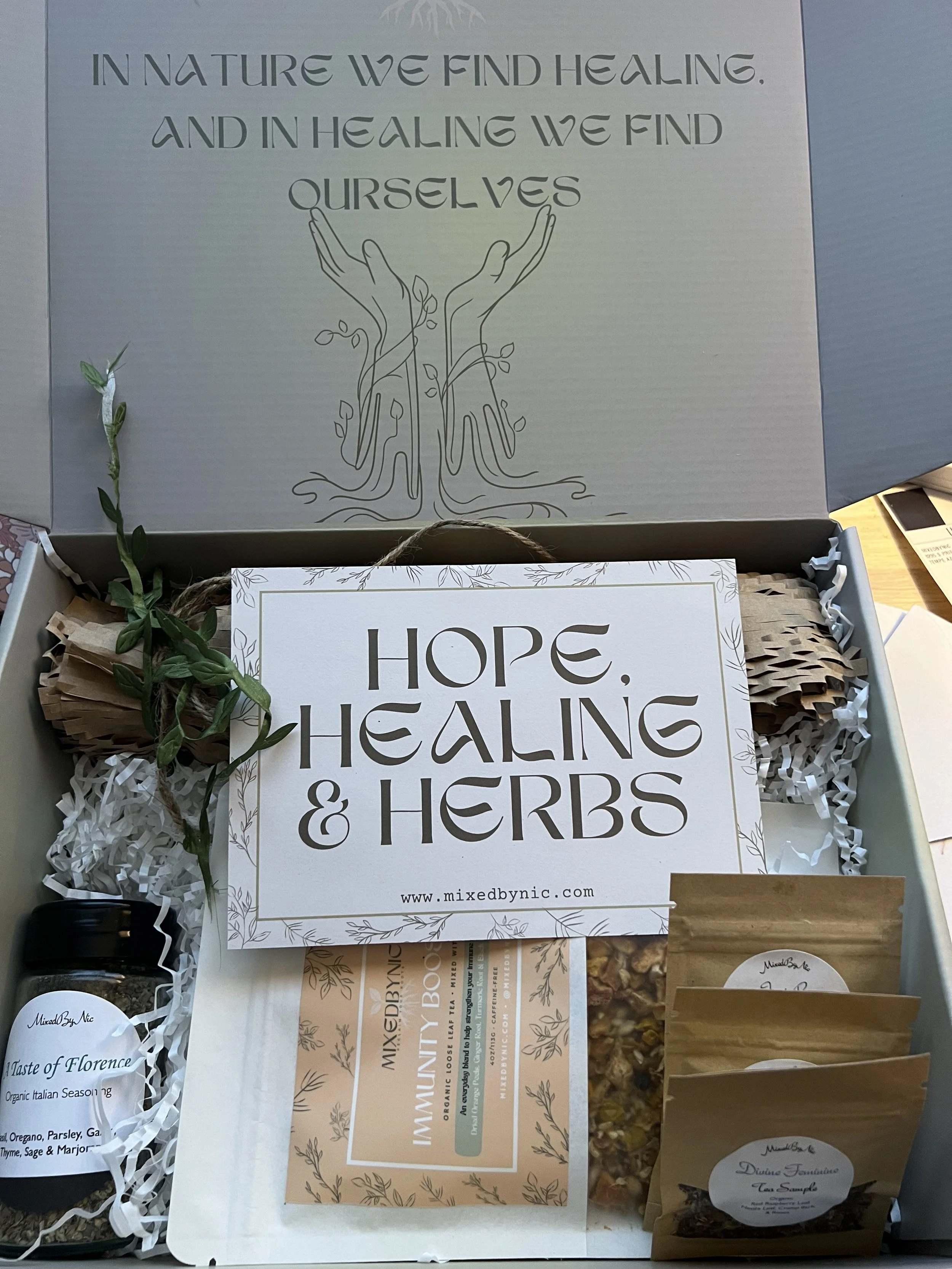 A wellness gift box filled with herbs, spices, and herbal blends, with a card reading 'Hope, Healing & Herbs,' and decorative paper, inside a box with a top flap featuring a quote about healing in nature and hands holding leaves.