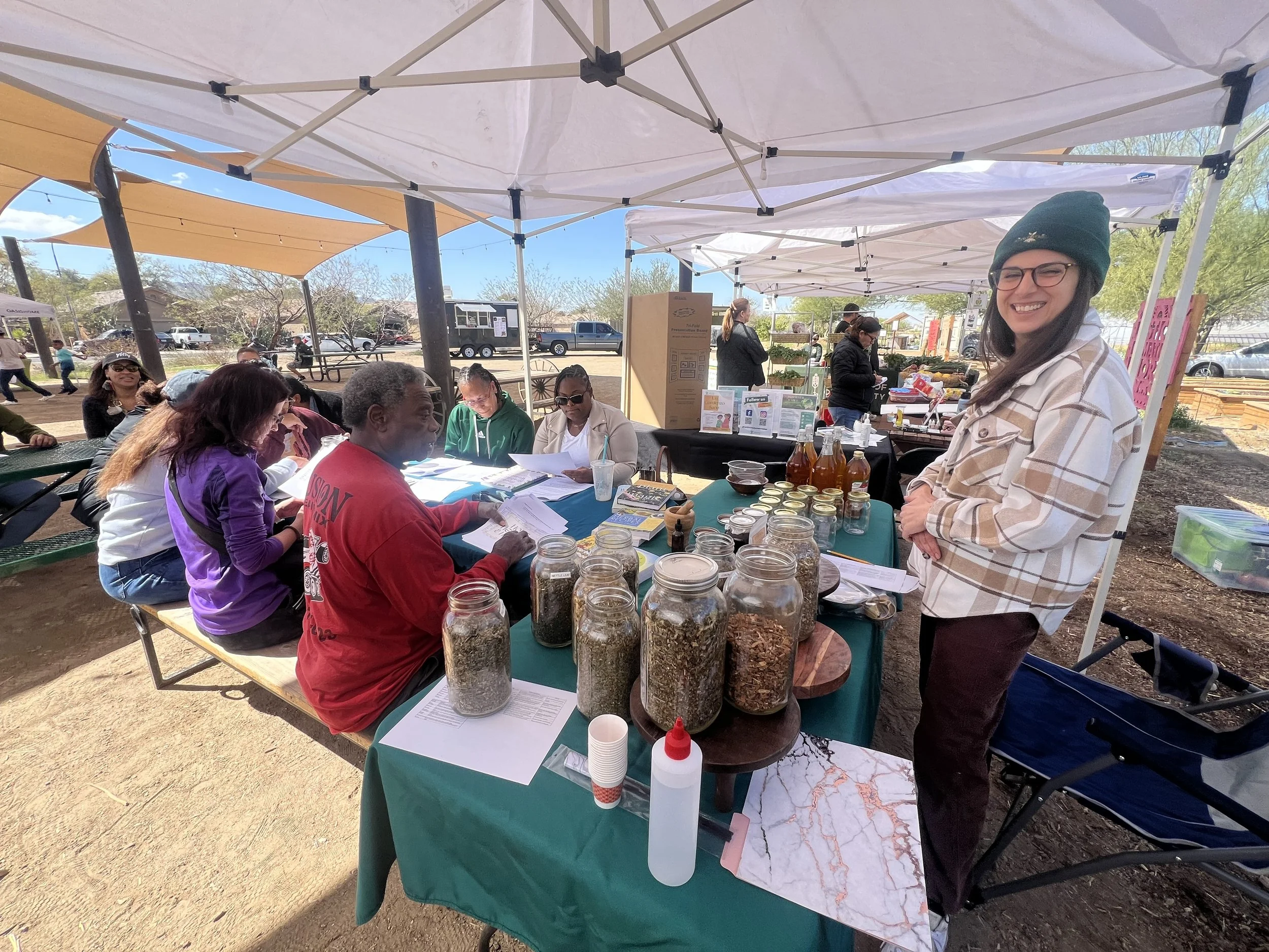 A woman in a green beanie and glasses stands smiling at a table with jars and bottles, while people sit and read papers under a white canopy at an outdoor market or event.