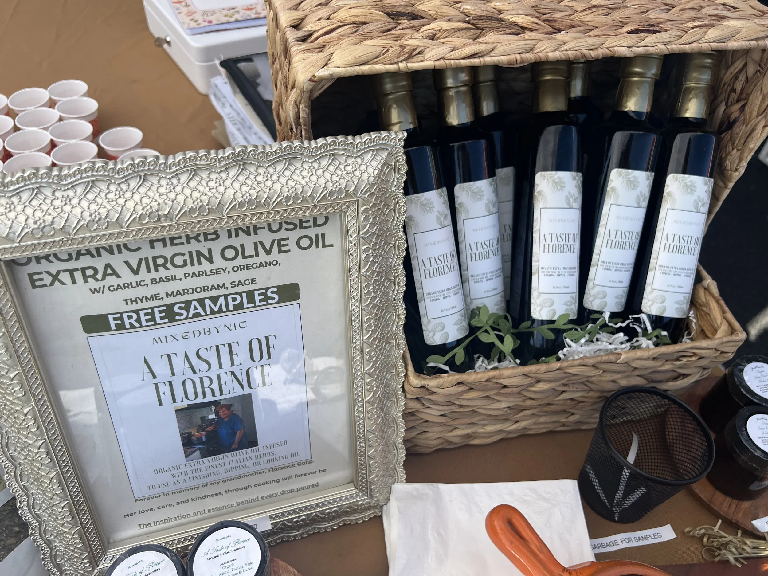Display of free samples of herbal-infused extra virgin olive oil labeled 'A Taste of Florence' in a basket. A decorative silver-framed sign details the herbal ingredients and the story behind the product.