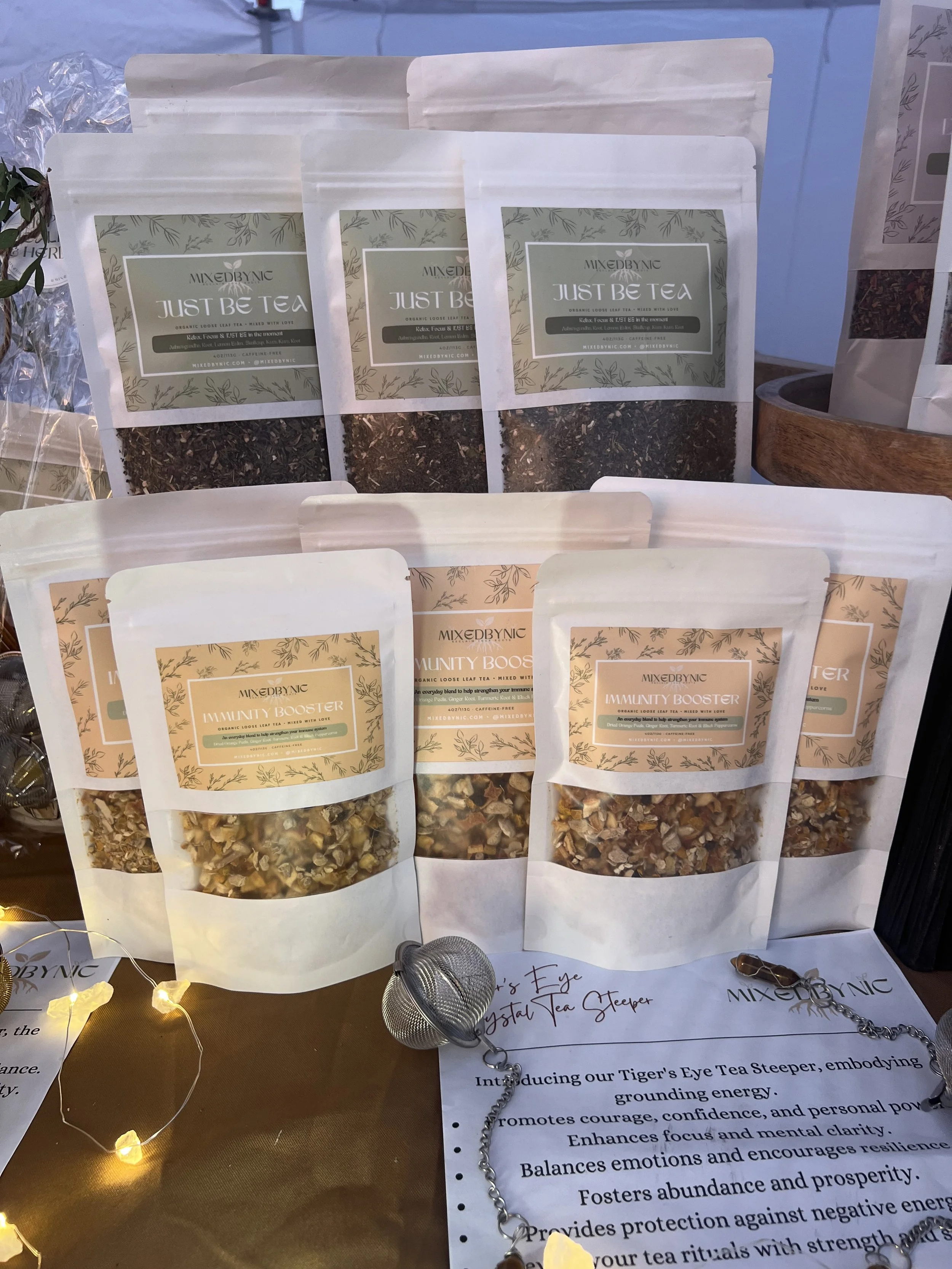 Display of tea products, including loose leaf tea and herbal tea blends, with packaging labeled "Just Be Tea" and "Immunity Booster." The setup includes a small tea infuser and a handwritten sign describing Tiger's Eye Crystal Tea Steeper, emphasizin