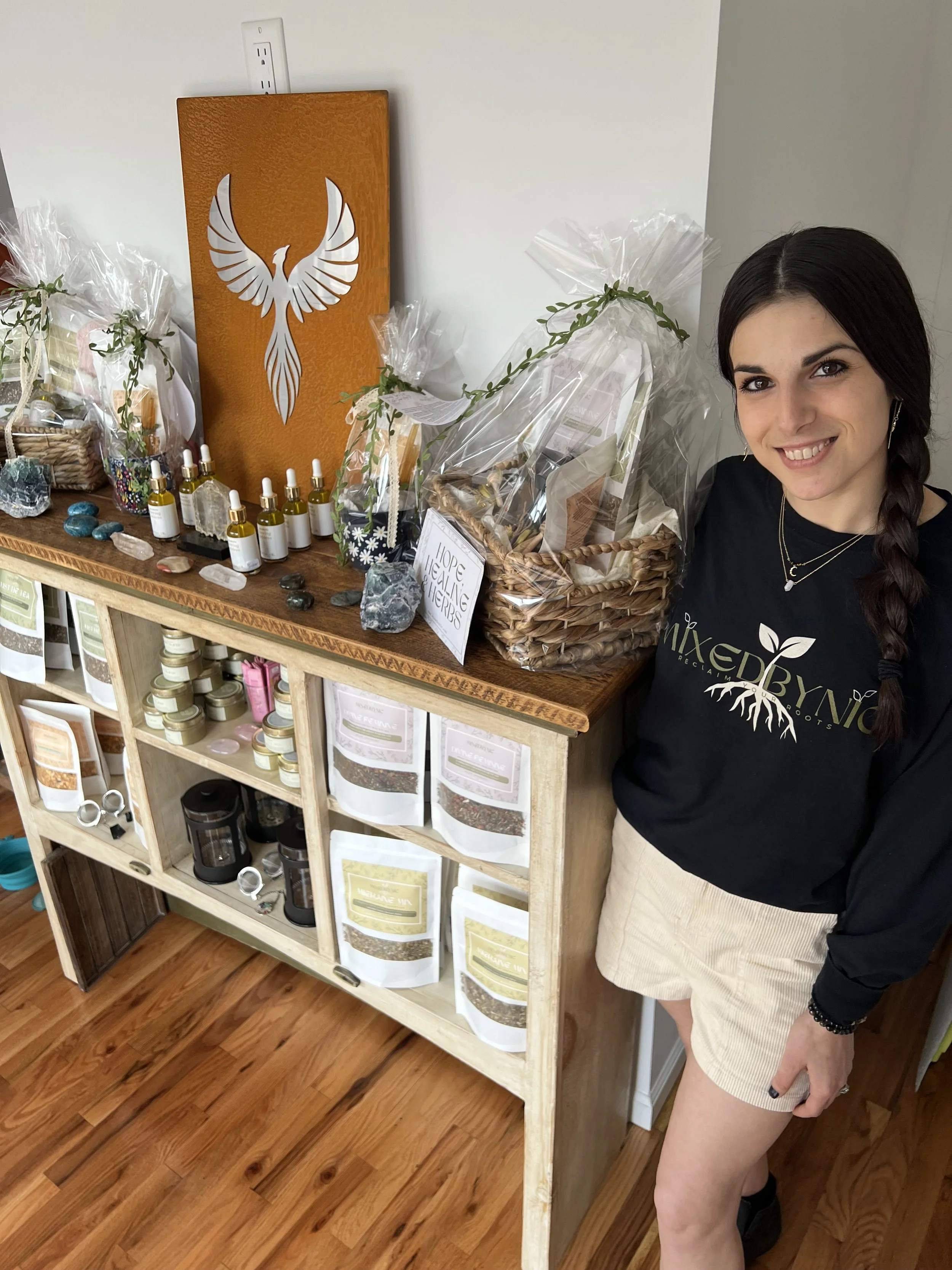 A smiling woman with dark hair in a braid, wearing a black sweatshirt and beige shorts, standing next to a display of essential oils, herbs, and wellness products on a wooden shelf and countertop.