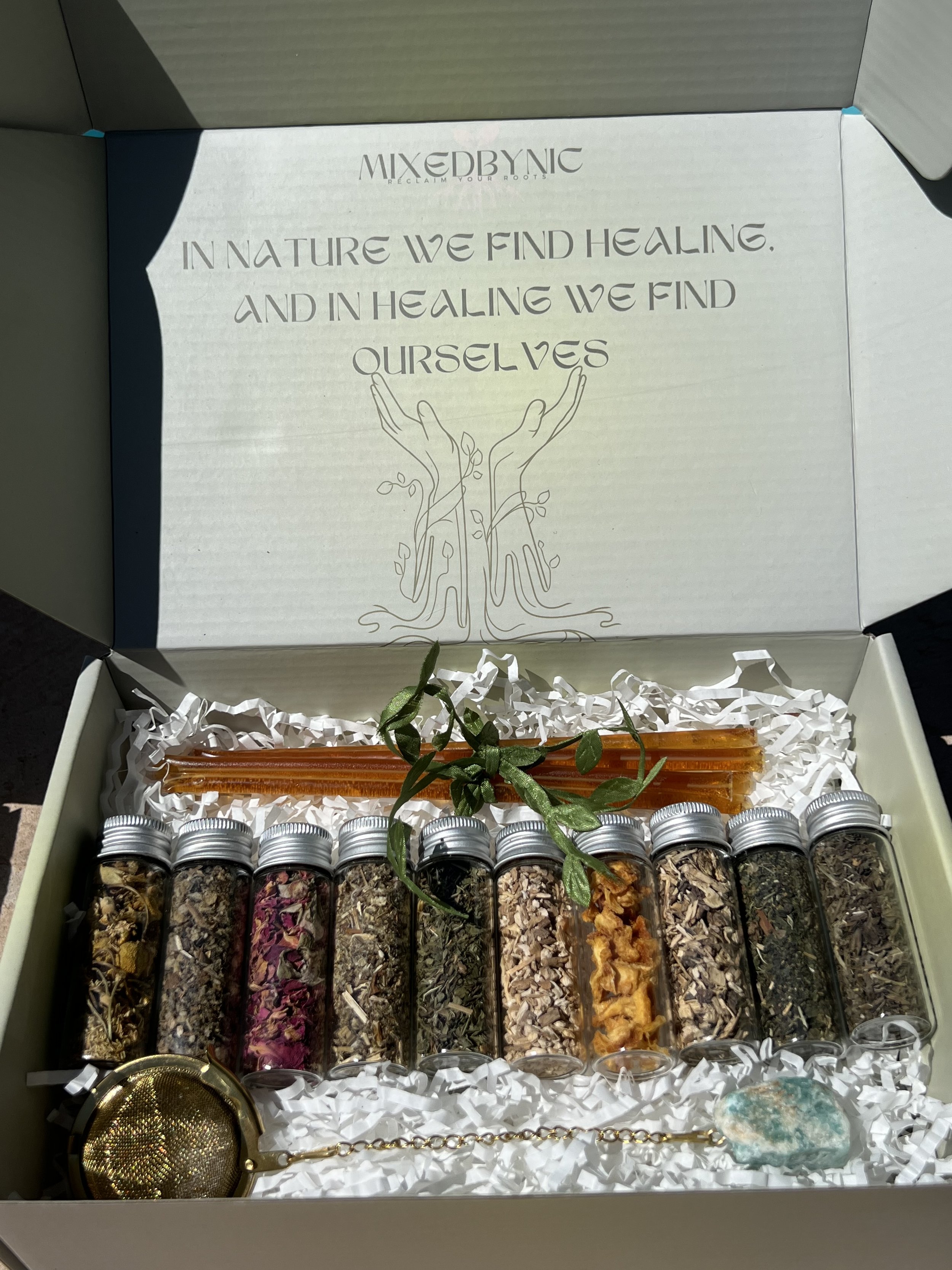 A wellness gift box containing various herbs and botanicals in small glass jars, a decorative spoon, a small plant, and an orange-colored resin stick, with a message about healing and nature on the inside of the box lid.