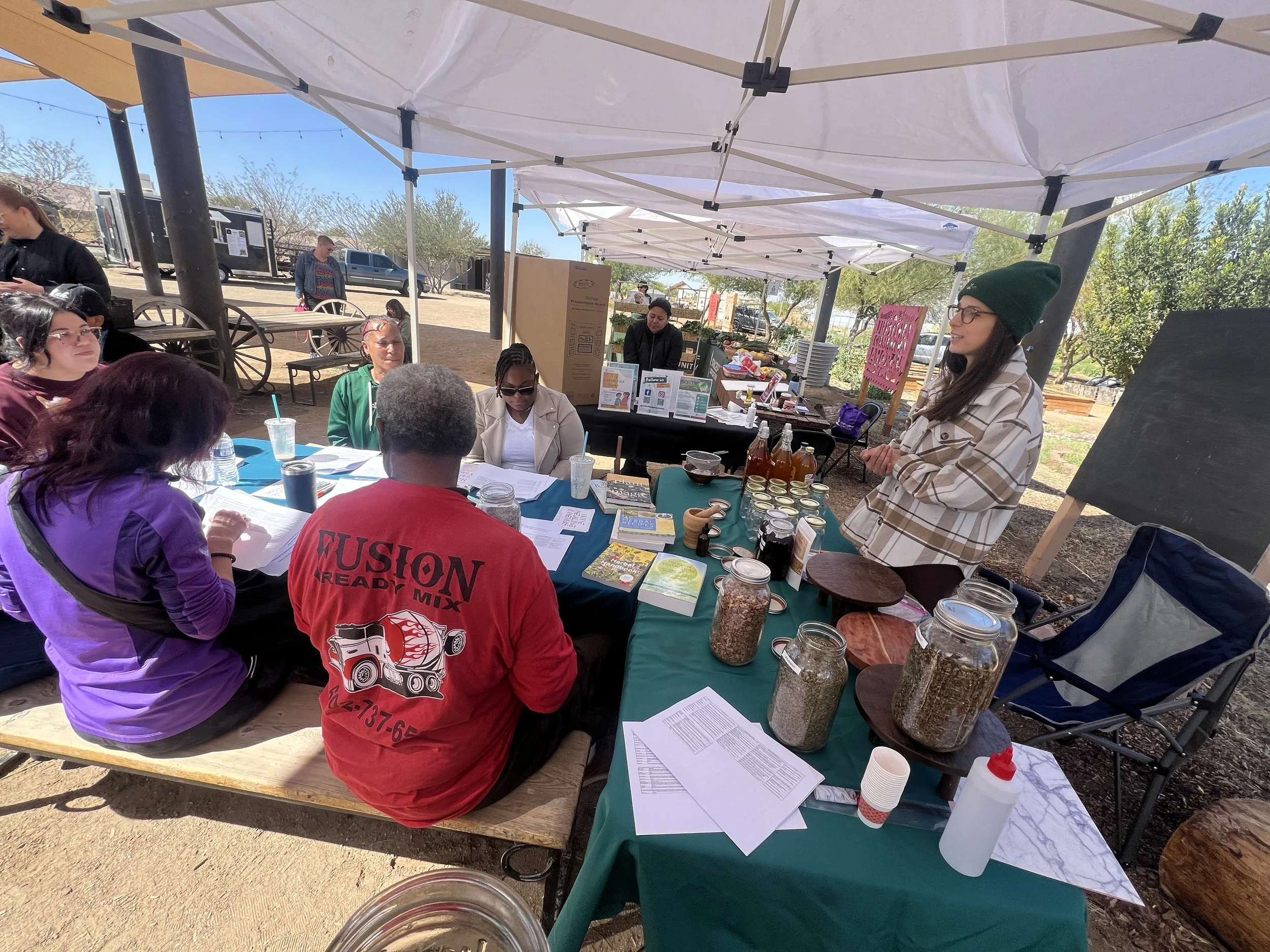 A group of people sitting at a table outside under a white canopy, participating in a workshop or discussion. The table has various jars, books, and papers. A woman stands on the right side, speaking to the group.