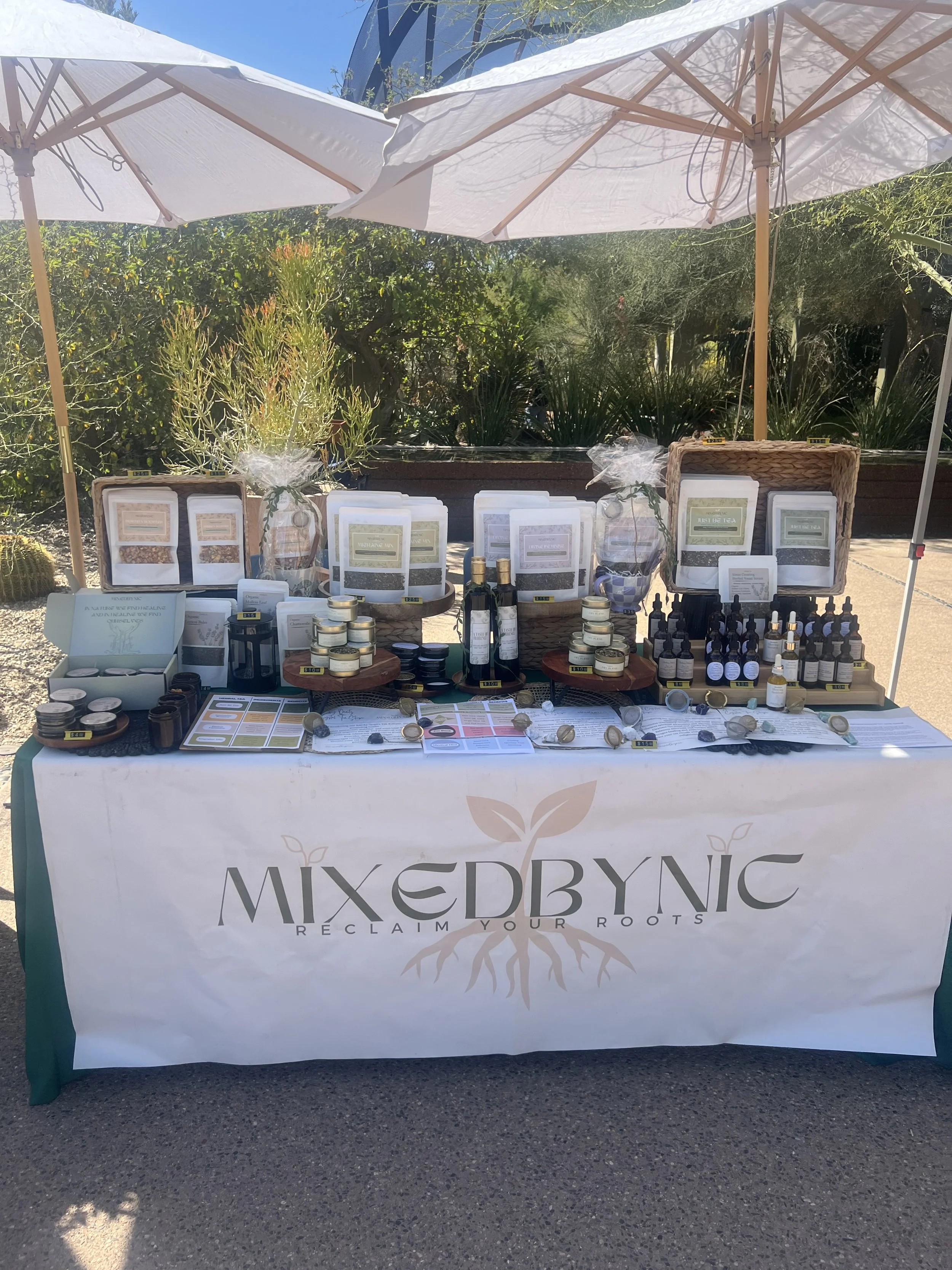Outdoor booth with herbal products and essential oils, branded as MIXEDBYNIC, under white umbrellas with a garden background.