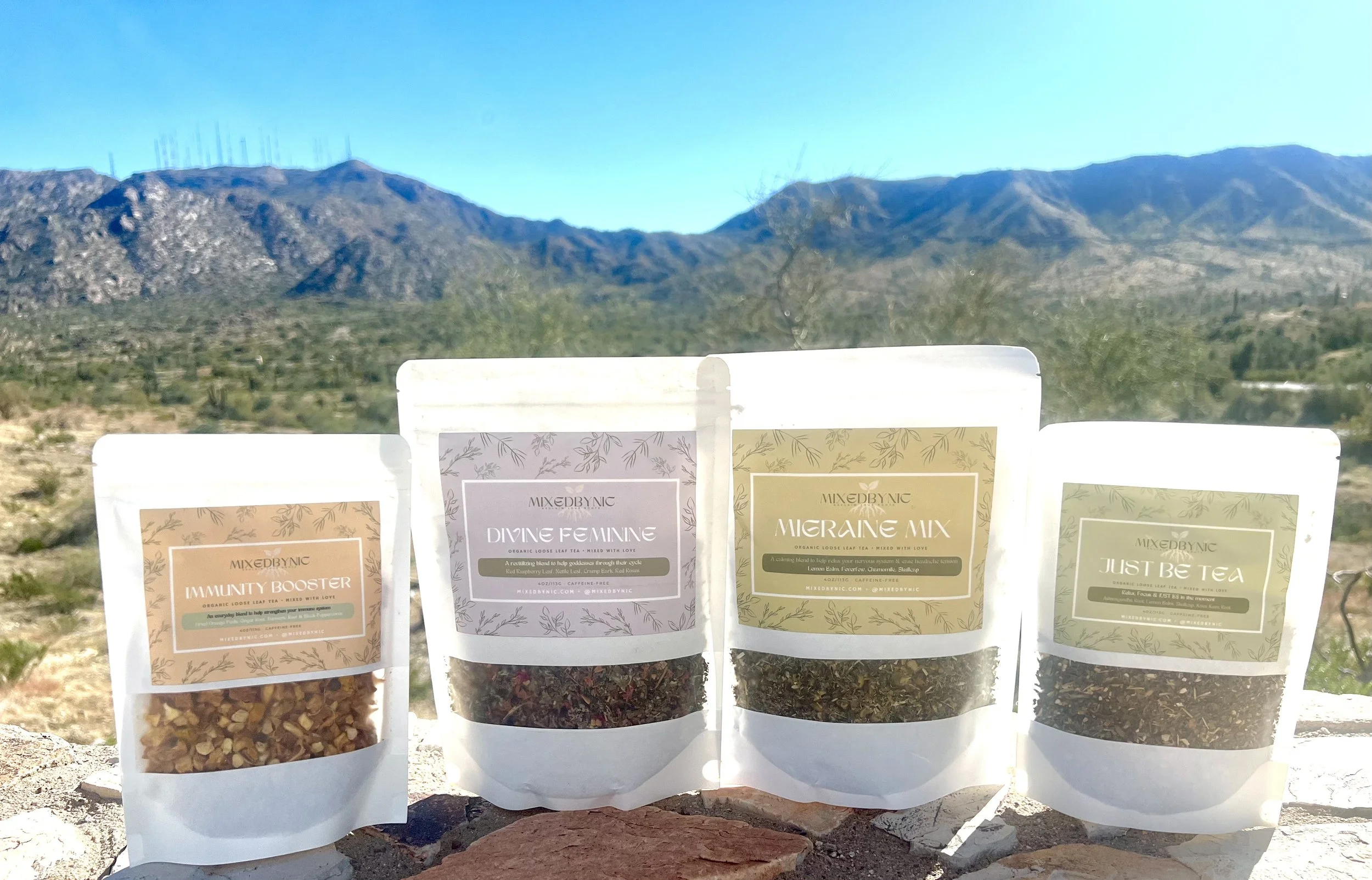Four herbal tea packages outdoors with mountains in the background. The packages are labeled 'Immunity Booster', 'Divine Feminine', 'Migraine Mix', and 'Just Be Tea'. The packages are standing on a ledge with rocky terrain and desert vegetation behin