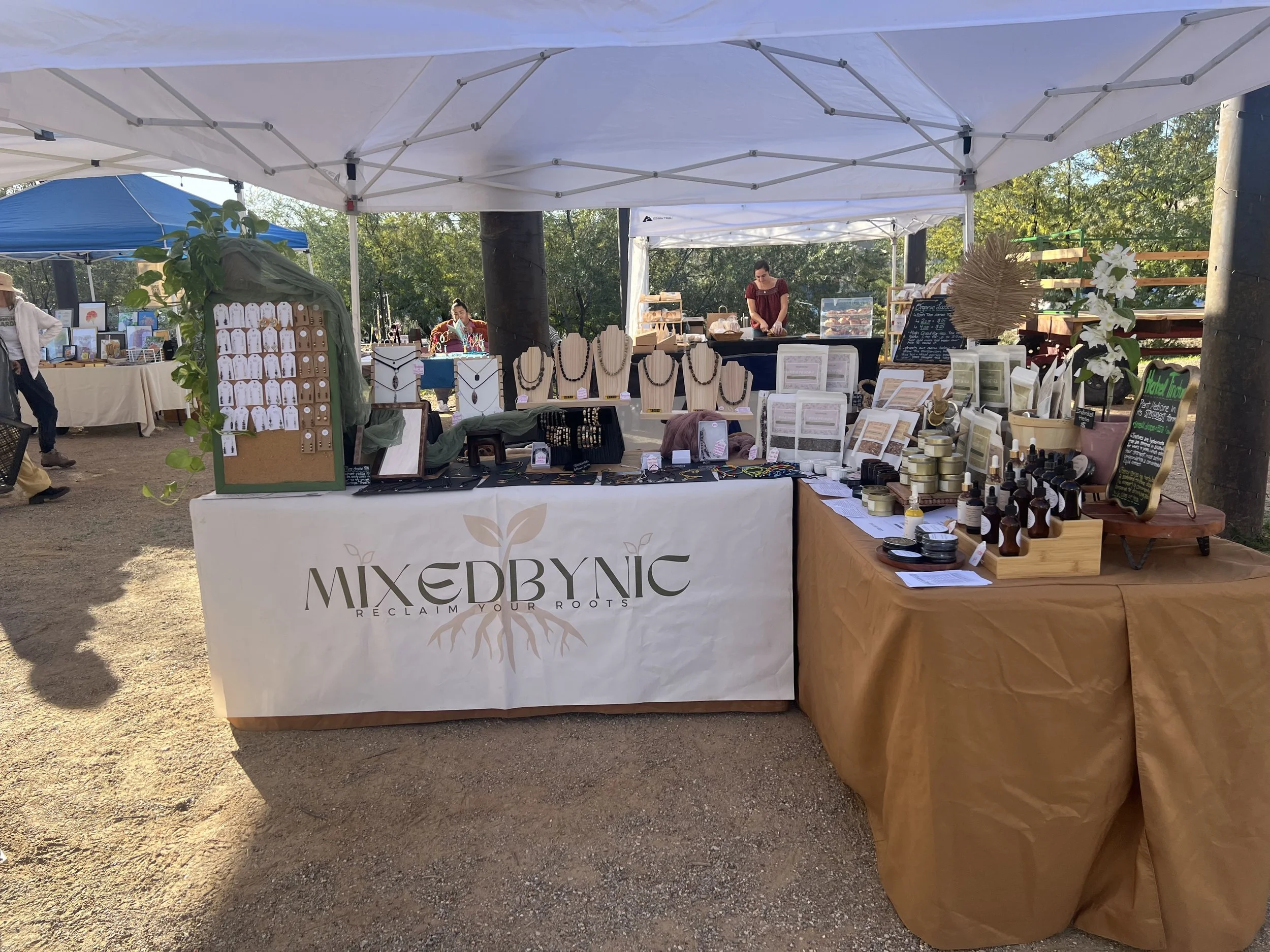 A market stall with jewelry and natural products on display at an outdoor event under white tents, with trees in the background.