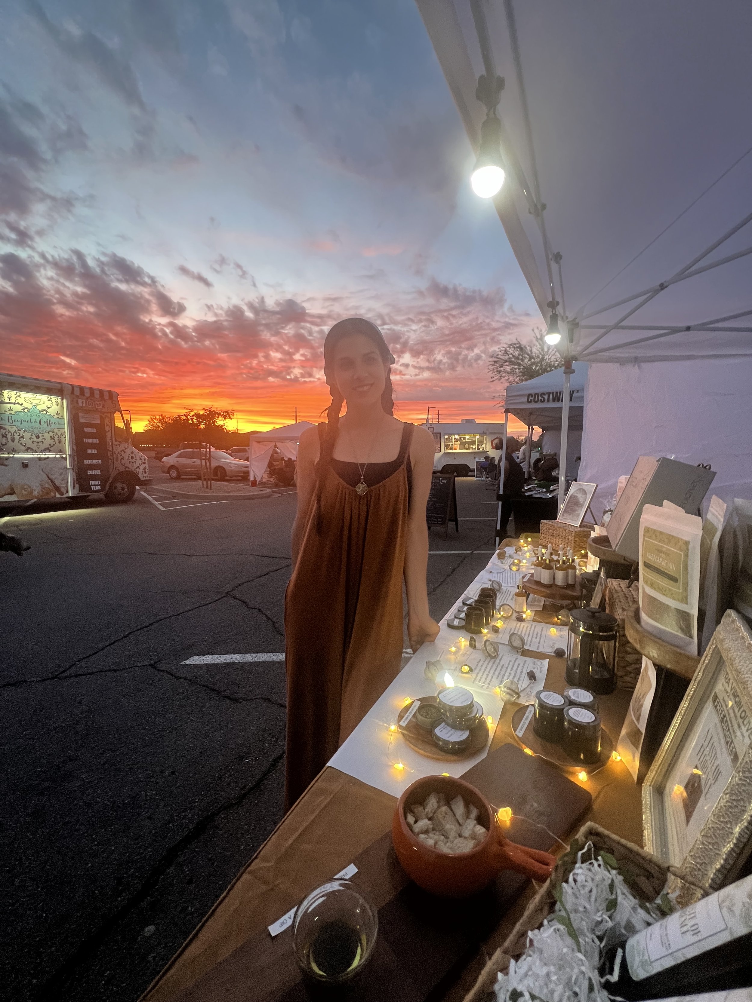 A woman standing next to a market stall at sunset, with tents and food trucks in the background, displaying small jars and framed pictures on the stall.