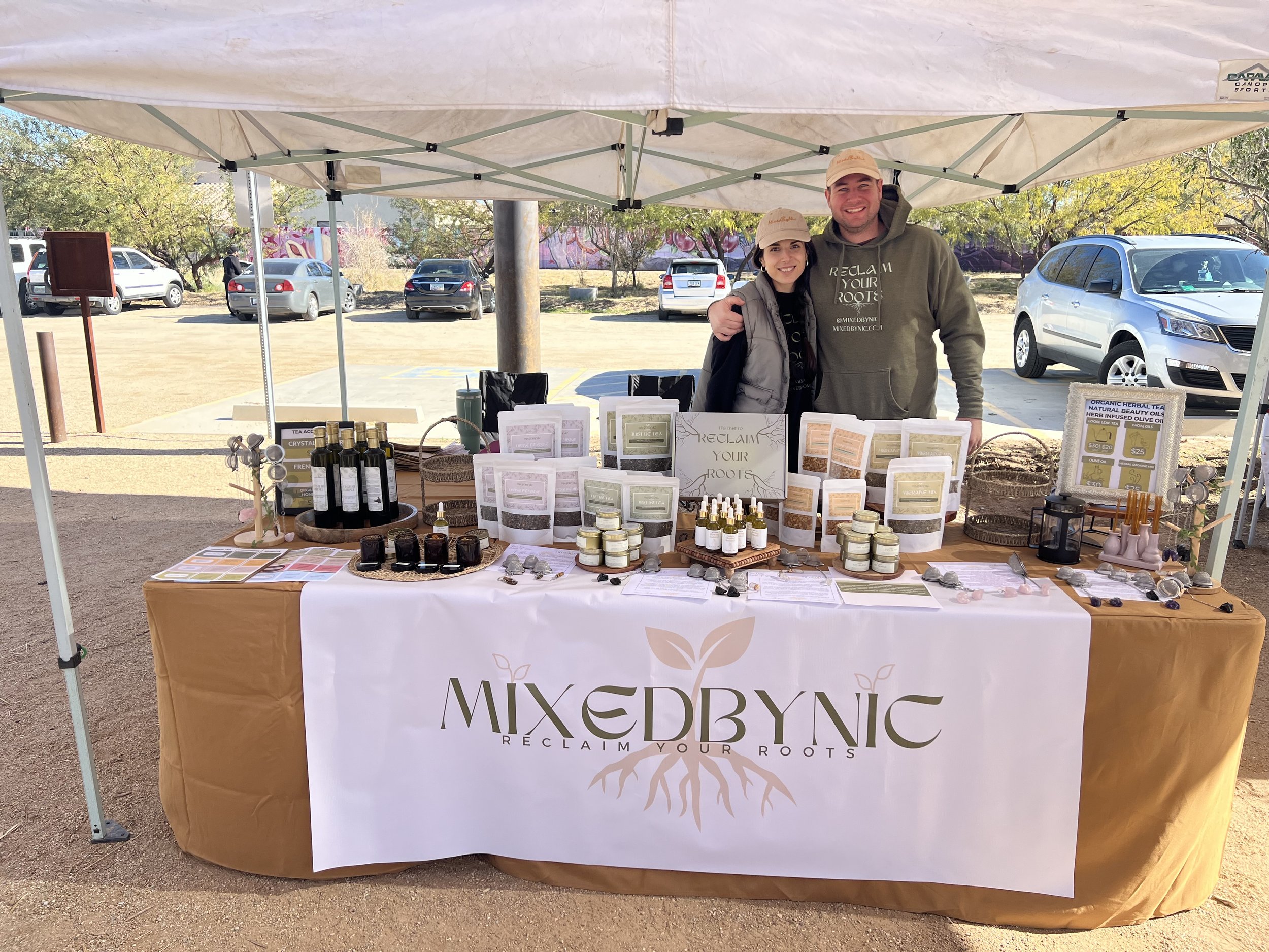 Two people standing behind a booth at an outdoor market, the booth displays various natural products, jars, bottles, and informational materials under a white canopy, with cars and trees in the background.