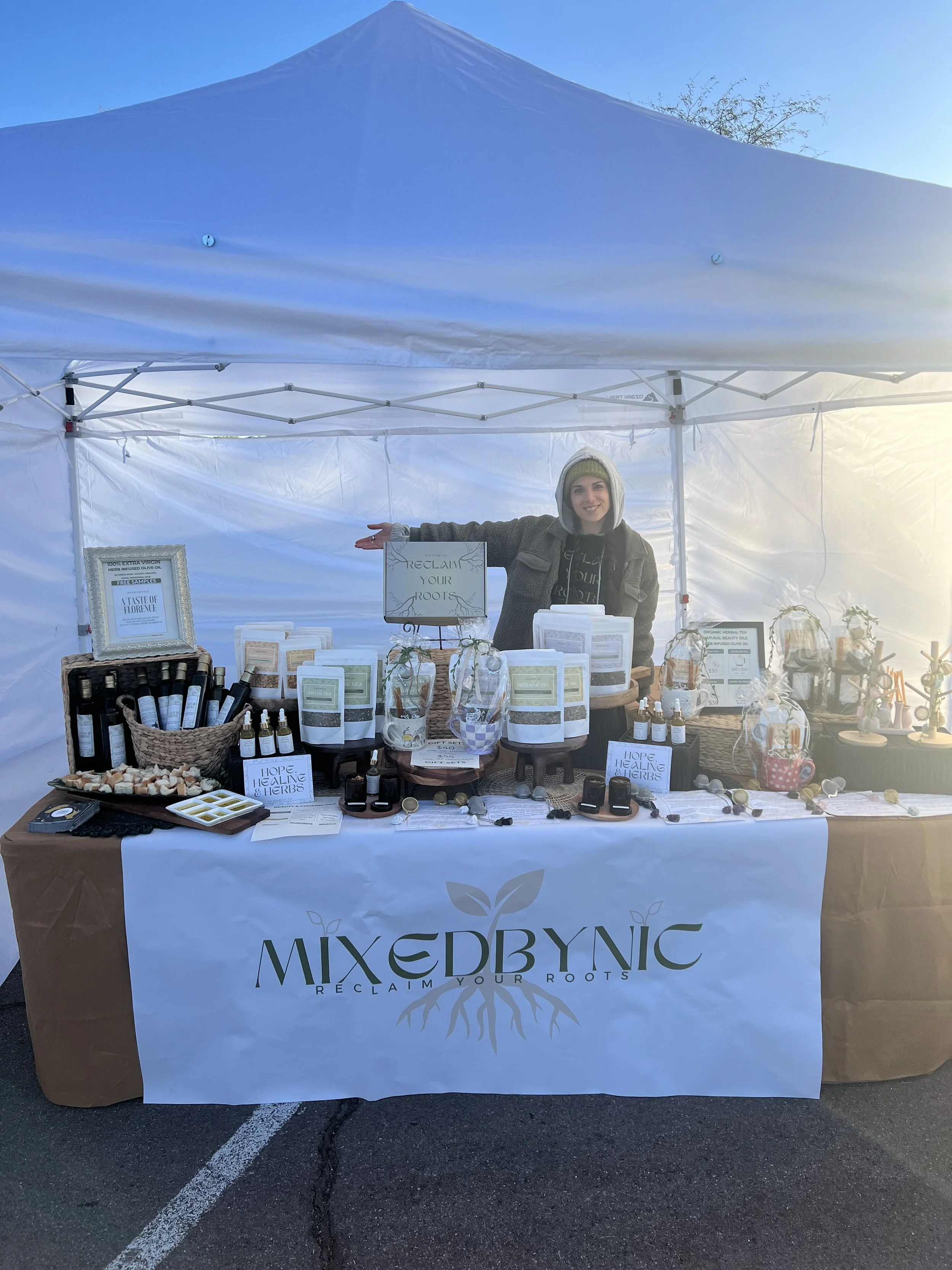 A woman at a booth selling herbal products under a white canopy tent. The table displays bottles, jars, and herbal packages, with signs reading 'Reclaim Your Roots' and 'Hope Healing & Herbs'. The booth has a banner with the logo 'MIXED BY NIC' and t