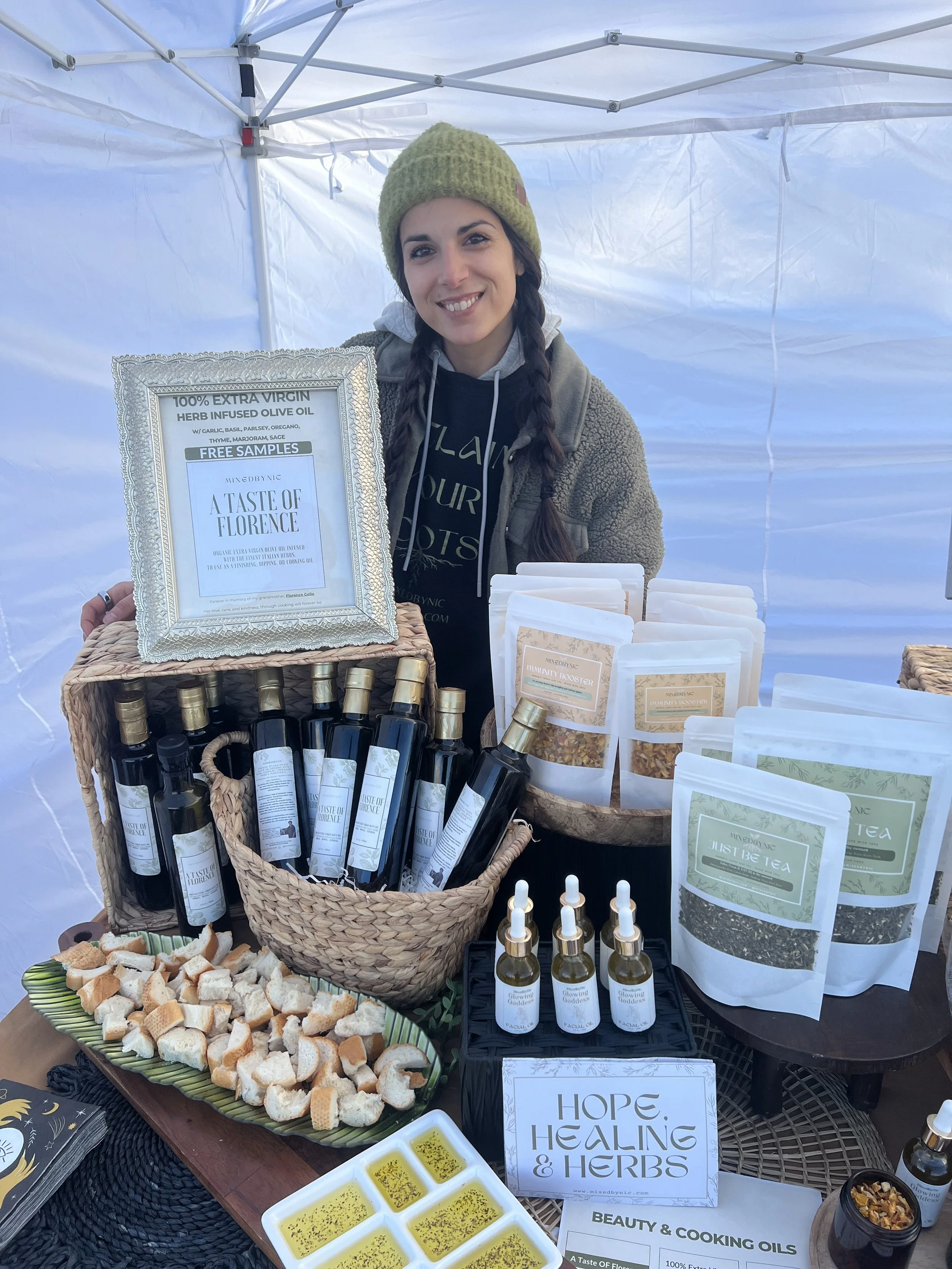 A woman standing behind a table at an outdoor market stall, displaying bottles of herbal olive oil, various herbal tea packages, and herbal remedies. She is smiling and wearing a green knit hat and a cozy jacket.