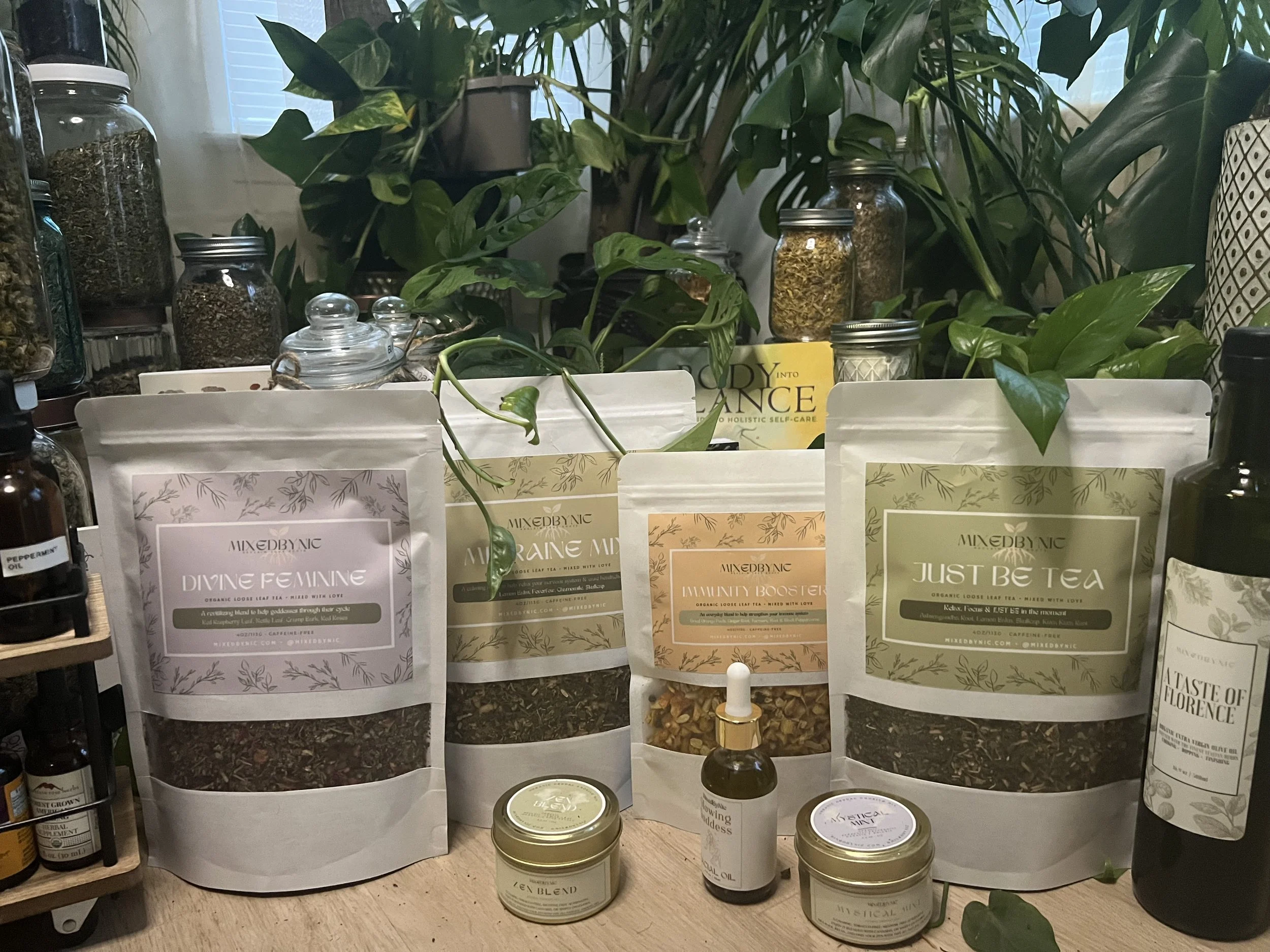 Assorted herbal teas and supplements in pouches and jars on a wooden surface surrounded by green houseplants.