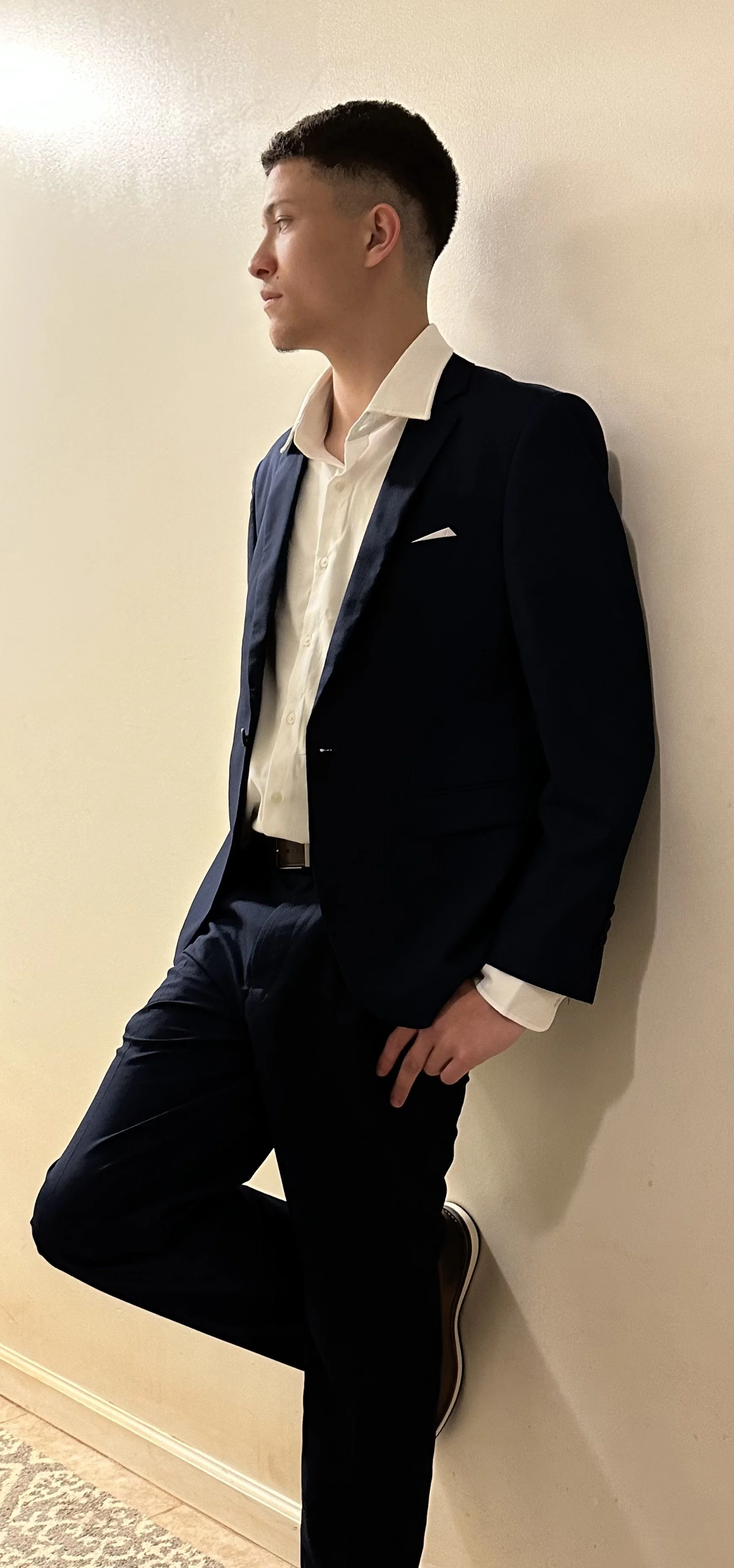 A young man in a navy blue suit and white dress shirt leaning against a wall, with one foot raised and resting on the wall.