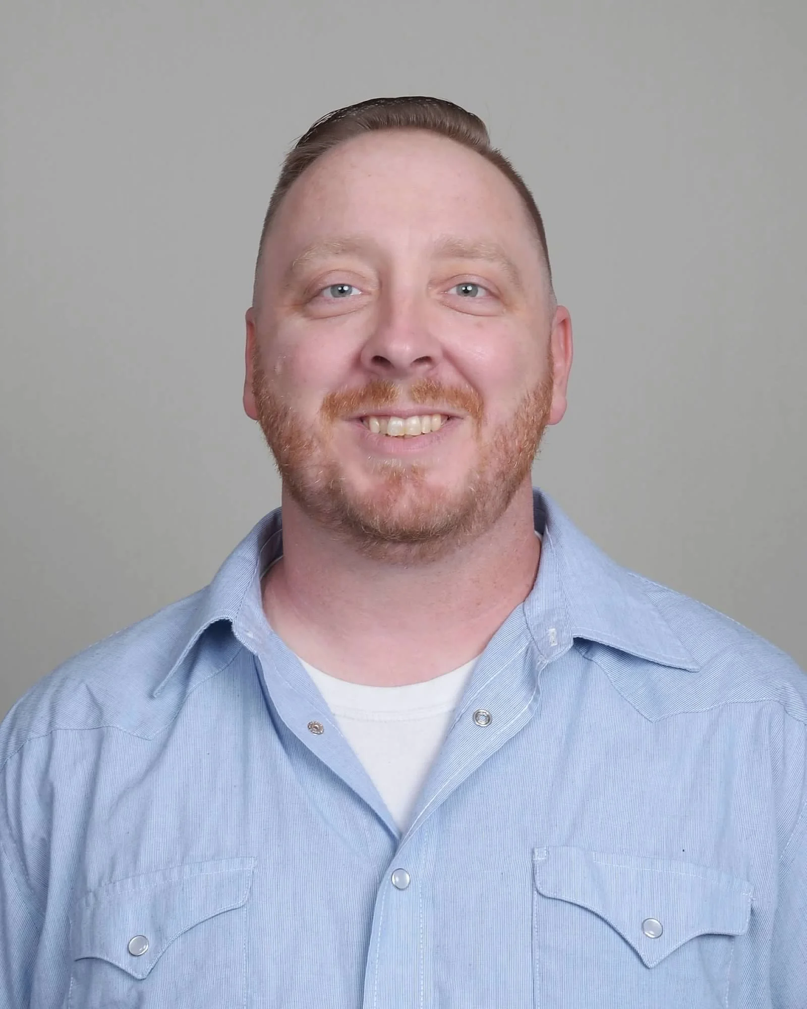 A smiling man with short brown hair, blue eyes, and a beard, wearing a light blue button-up shirt against a neutral gray background.