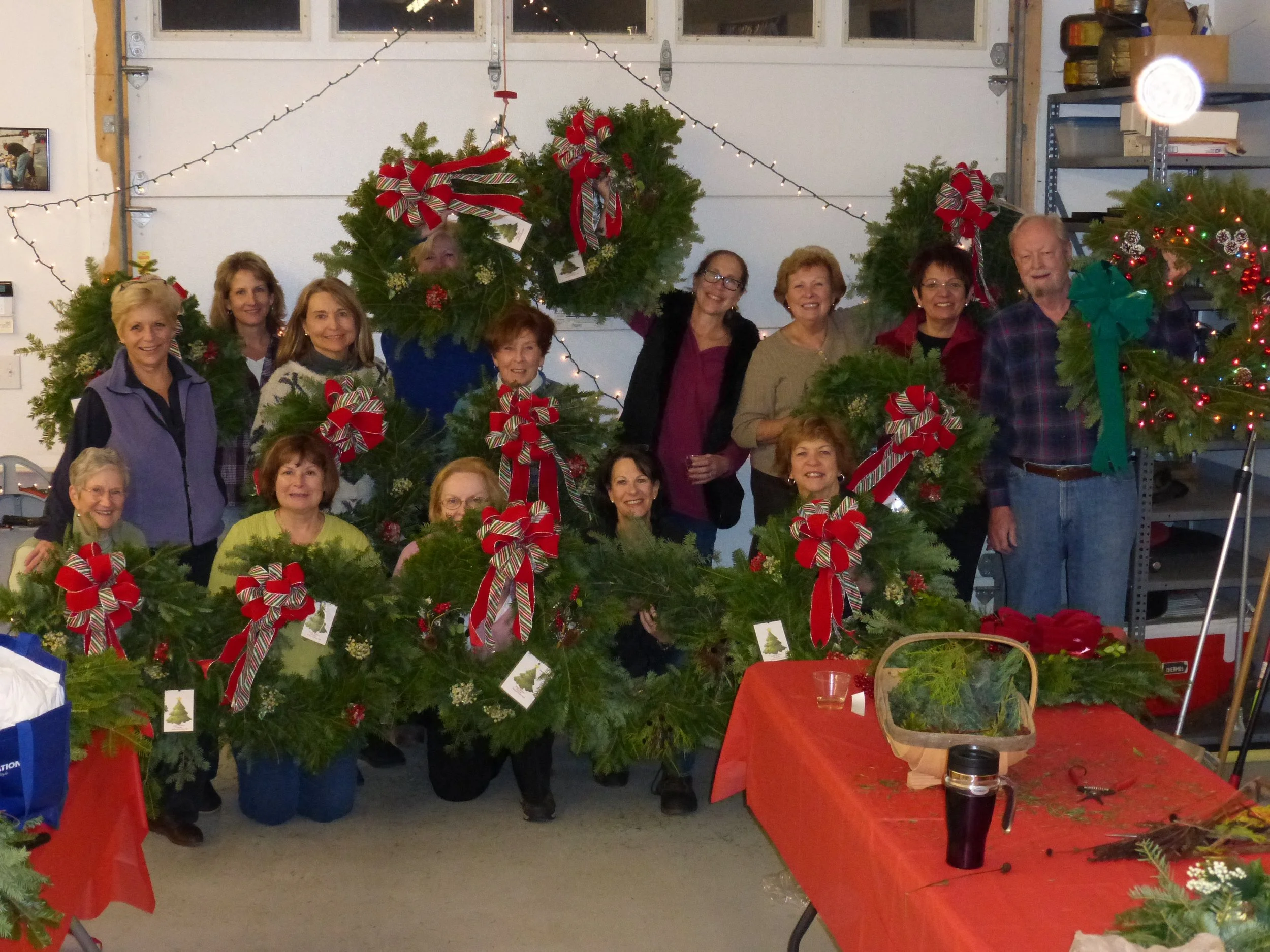 Group of people in a garage, posing with decorated holiday wreaths featuring red and green ribbons, holiday ornaments, and greenery.