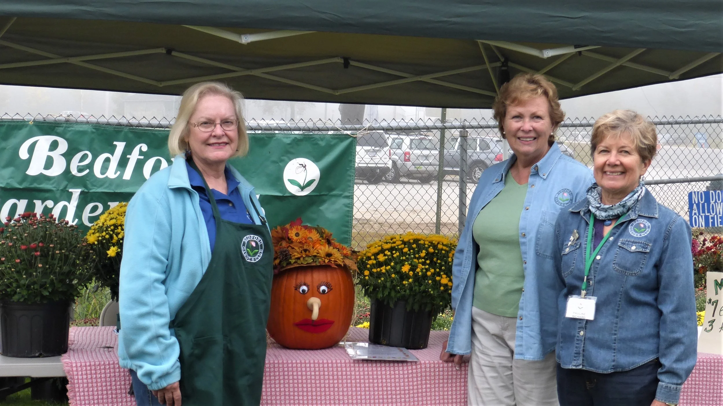 Three women standing in front of a table with potted chrysanthemums and a decorated pumpkin with a face, under a green canopy at Bedford Garden event.