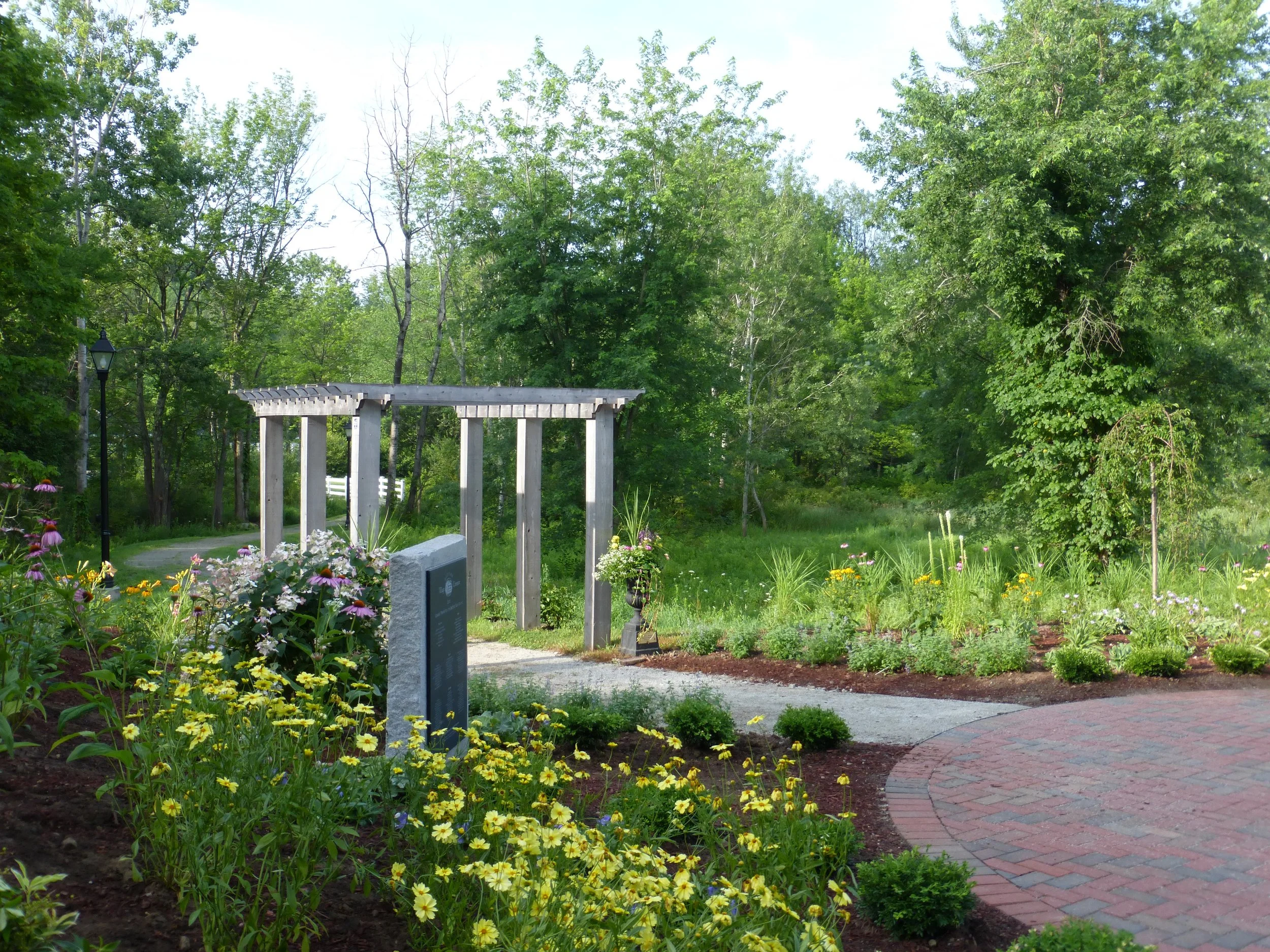 A landscaped garden with a brick pathway, colorful flowers, a pergola, a lamppost, and lush green trees in the background.