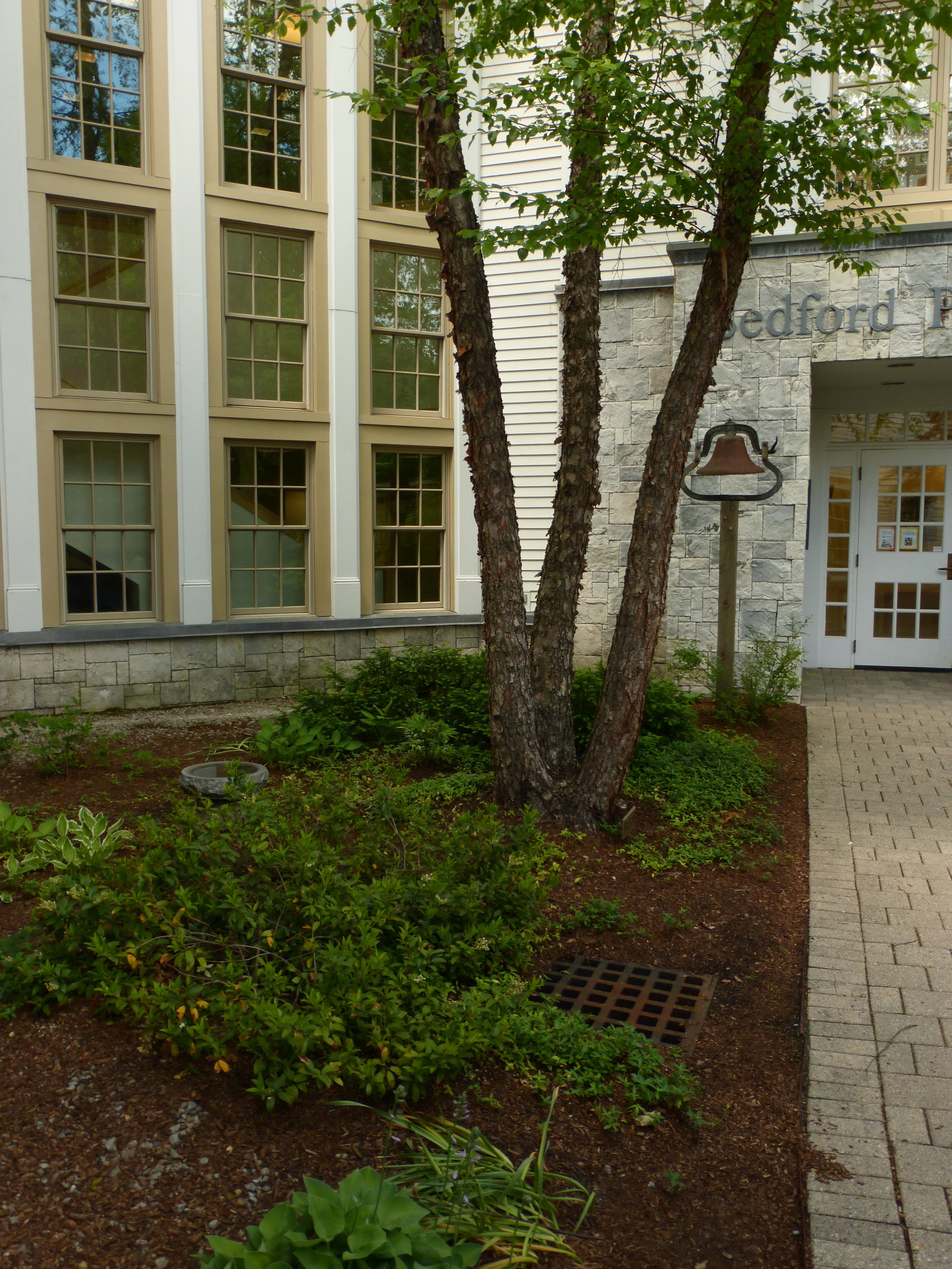 Exterior of a building with a stone and siding facade, multiple windows, a sign that reads 'Bedford P,' a lamp post, and a tree surrounded by shrubs and a paved walkway.