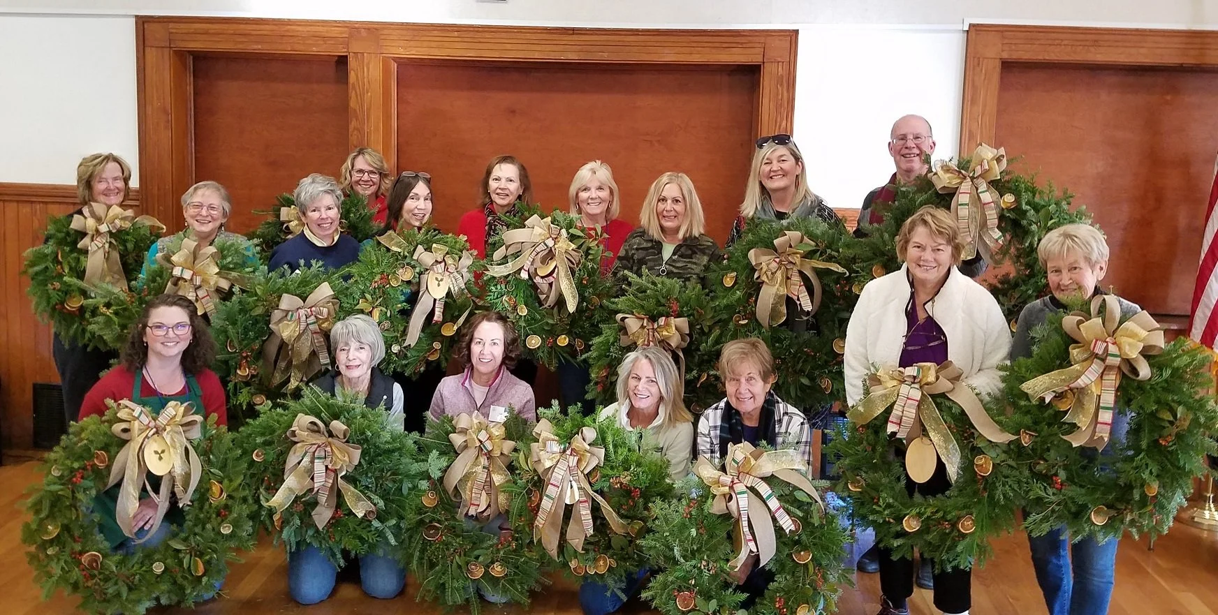 A group of women and one man posing indoors with decorated Christmas wreaths, all smiling and holding wreaths adorned with gold ribbons, ornaments, and greenery.