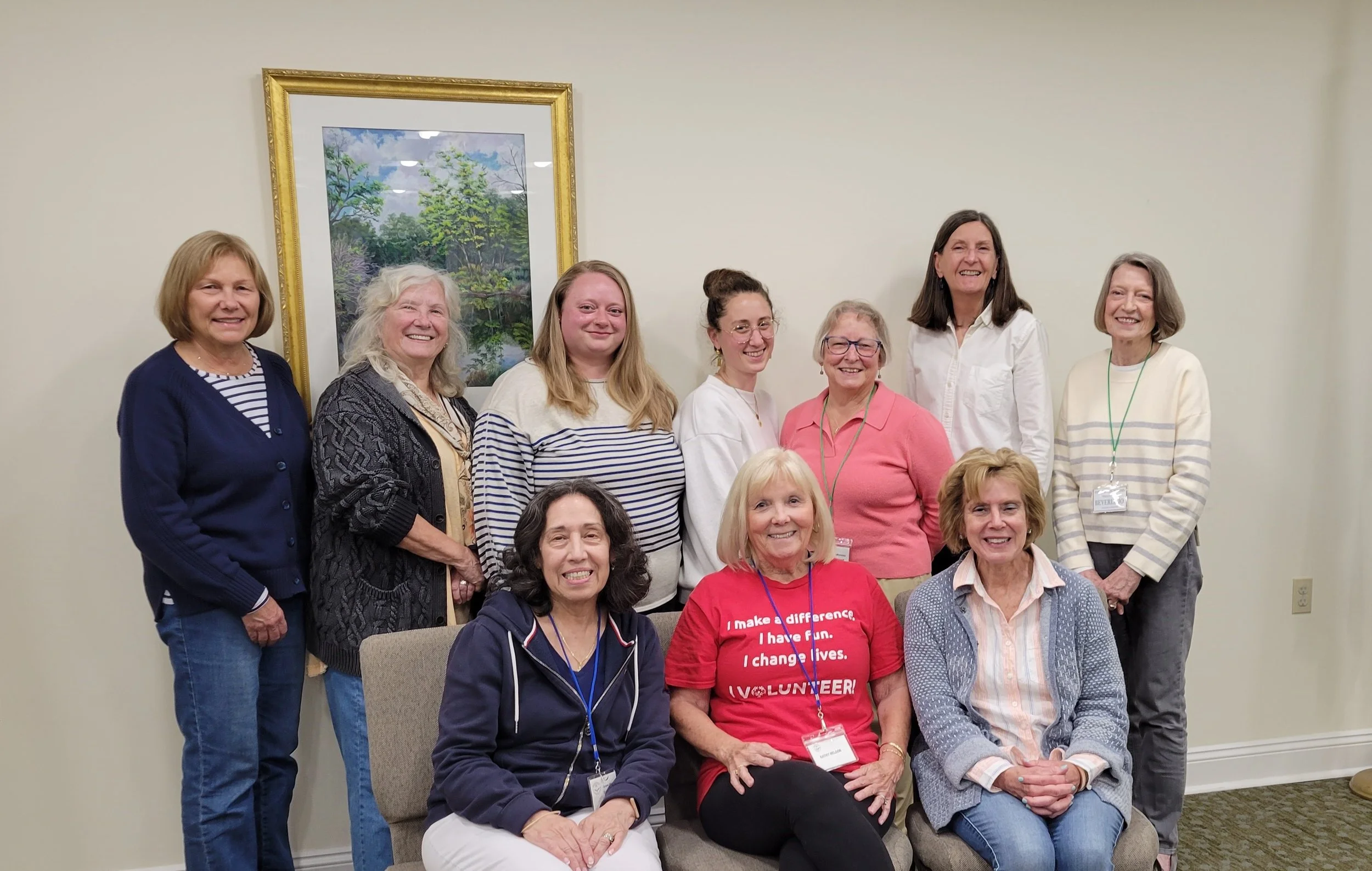 A group of ten women, some seated and some standing, smiling for a photo in an indoor setting. There is a painting of trees on the wall behind them, and they are wearing casual attire, some with name badges.