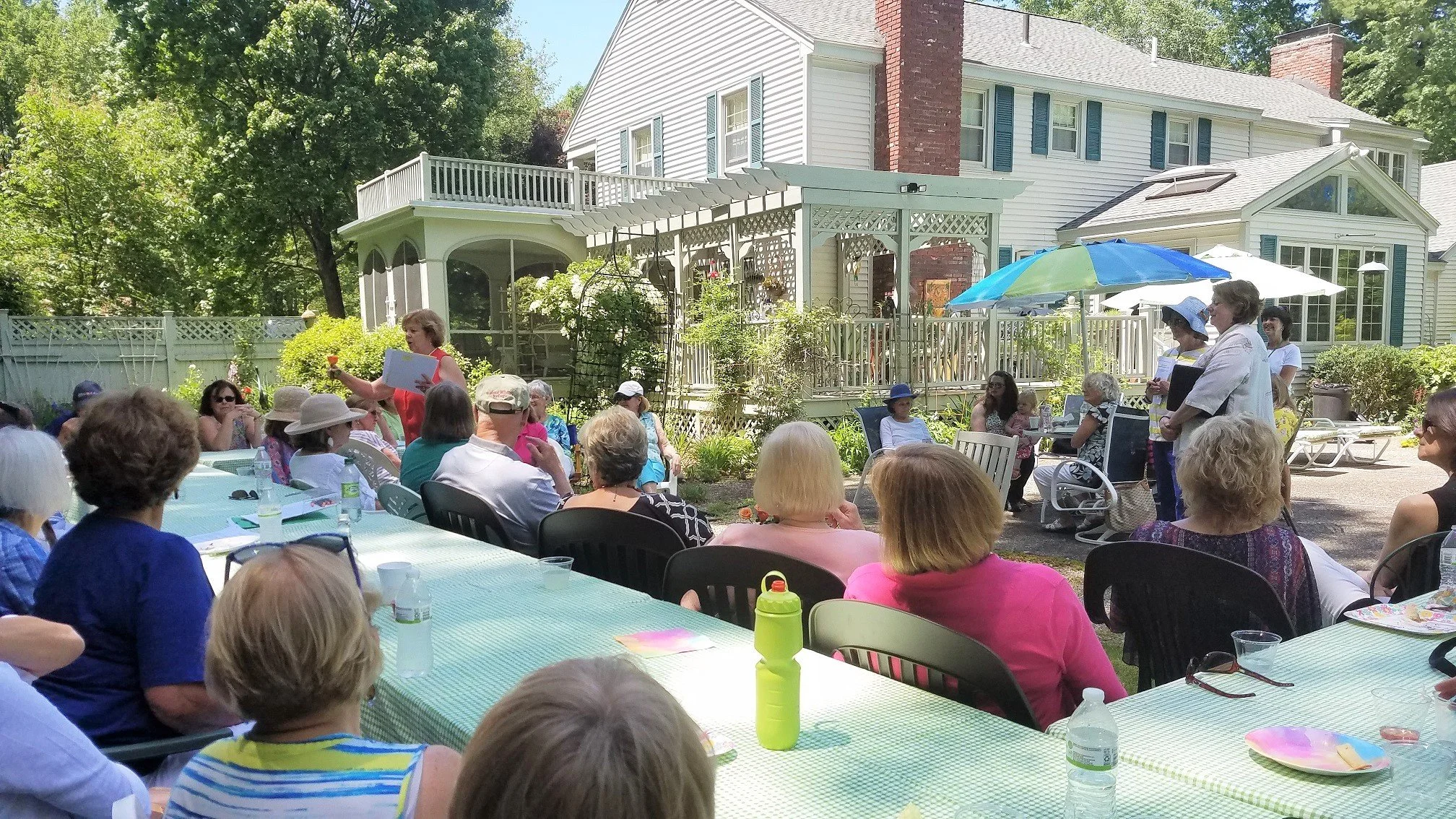Outdoor gathering of mostly women seated at long tables in a backyard, with a woman standing and speaking to the group. The background features a large white house with a deck, trees, and umbrellas providing shade.