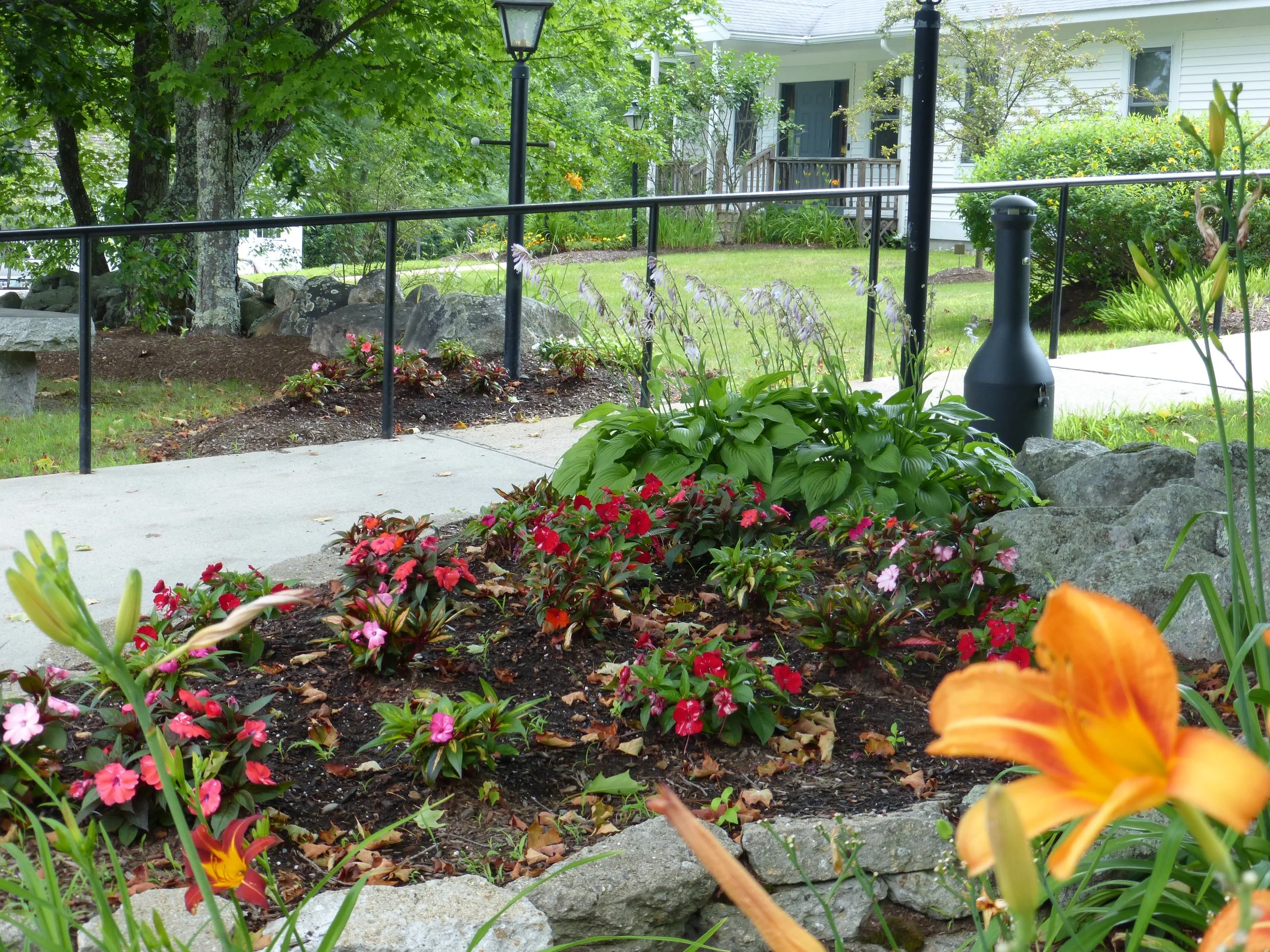 A landscaped garden with colorful flowers, green plants, rocks, a concrete pathway, and a black lamp post in front of a white house with a porch and a black railing.